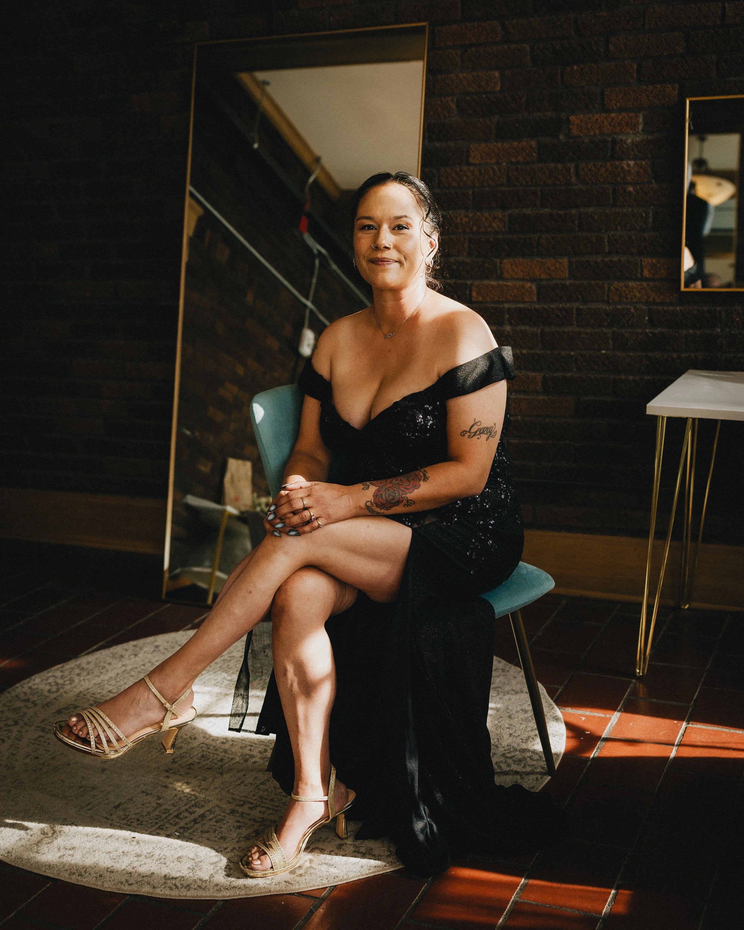 A woman in a black evening dress and heels sitting on a teal chair in a dimly lit room, with dark brick walls and a large mirror behind her. Shot at Topeka Kansas venue The Beacon.