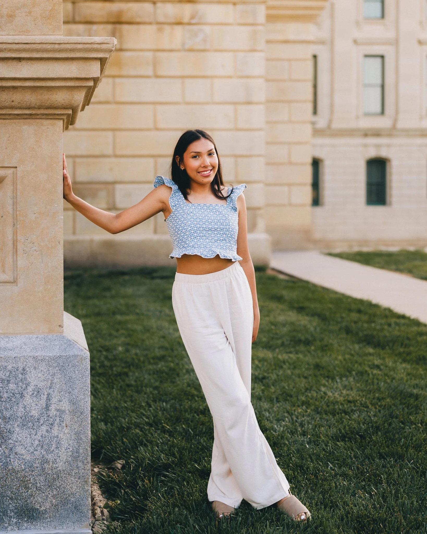 A young woman stands outdoors on grass, leaning against a stone building. She is wearing a blue floral crop top with ruffled sleeves and white wide-leg pants, smiling at the camera.