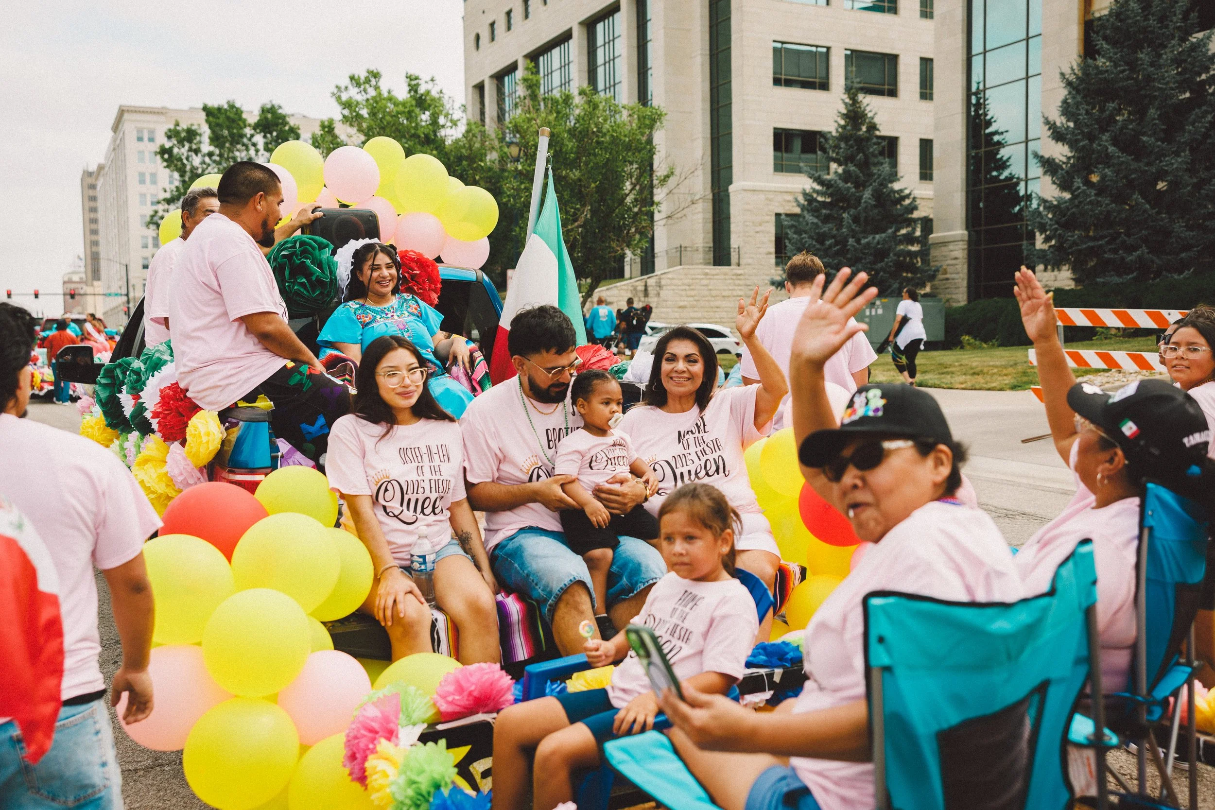 Group of diverse people celebrating at a parade, riding a colorful decorated float with balloons, flowers, and Mexican flags, with city buildings in the background. Shot at Fiesta Topeka parade.