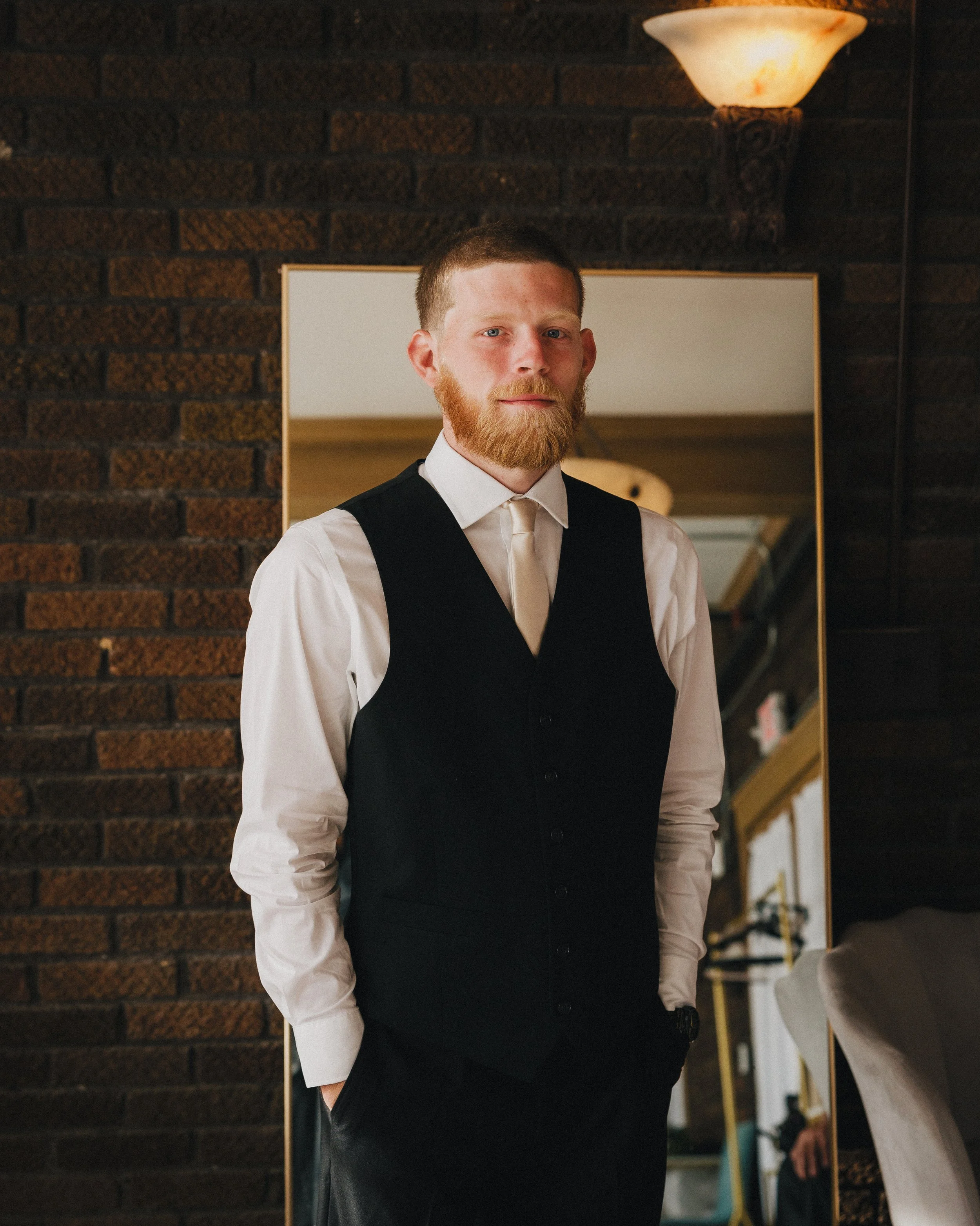 A man with a beard and short hair standing indoors in front of a mirror. He is wearing a white shirt and a black vest, with a serious expression. The background includes a brick wall and a wall-mounted lamp. Shot at Topeka Kansas venue The Beacon.