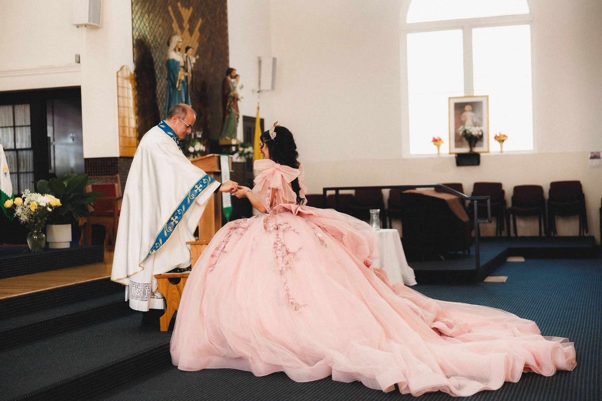 A young woman in a pink quinceañera dress receiving a blessing from a priest during a religious ceremony in a church.