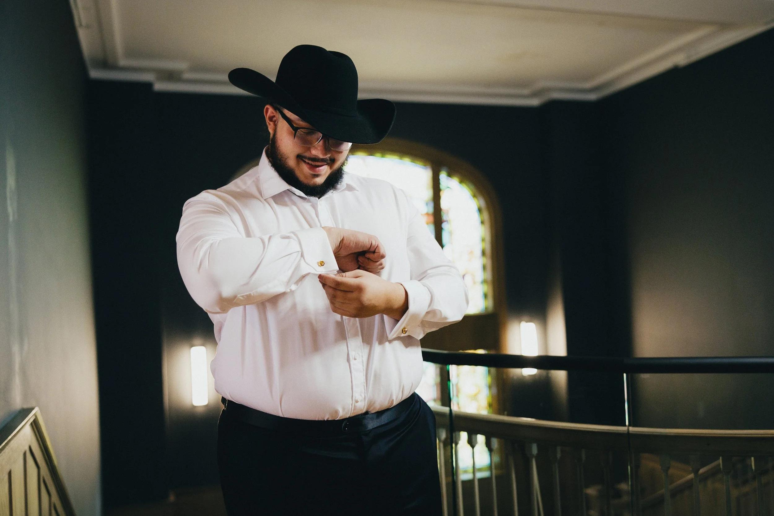 A man wearing a black cowboy hat, glasses, and a white dress shirt is smiling as he looks down and adjusts his cufflinks inside a building with a stained glass window in the background. Shot at Topeka Kansas venue The Beacon.
