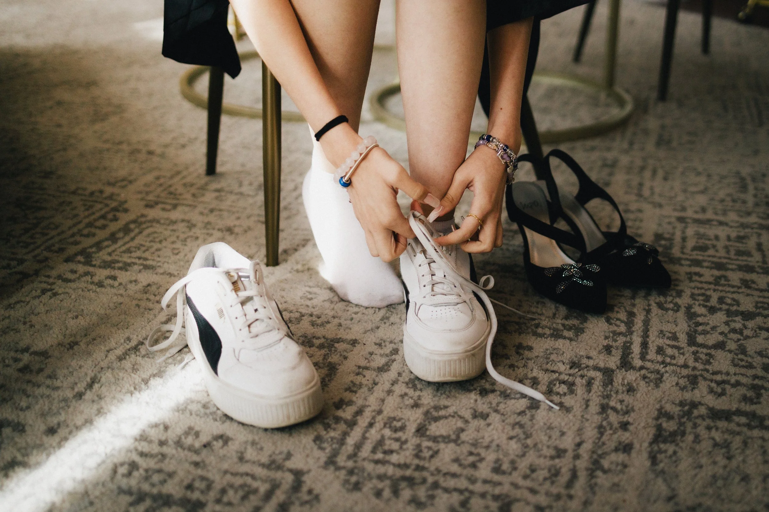 Person tying their white sneakers while sitting on a chair, with black high-heeled shoes beside them on a patterned carpet. Shot at Topeka Kansas venue The Beacon.