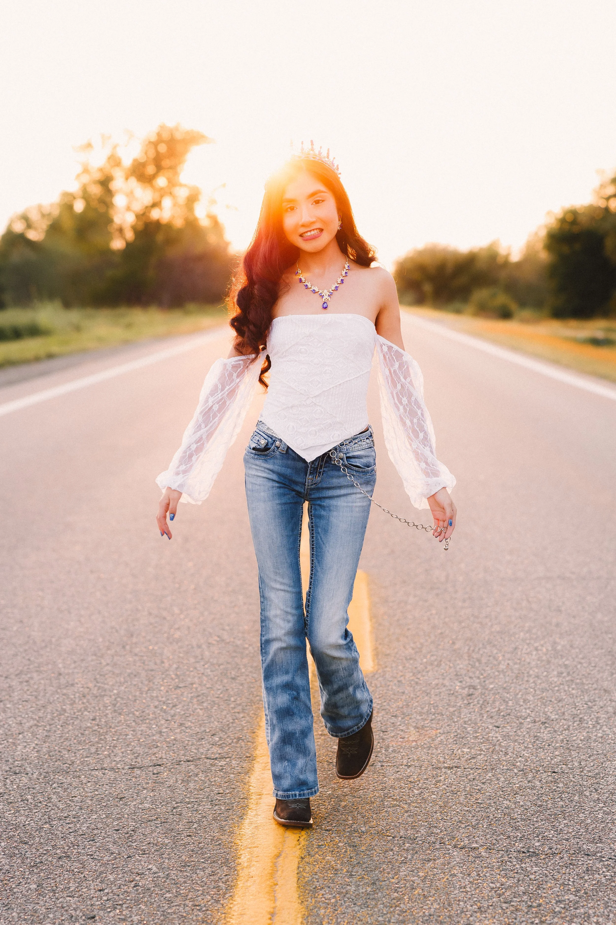 A young woman with long dark hair walking on a road during sunset, wearing a white off-shoulder top, jeans, cowboy boots, and jewelry, with a crown on her head.