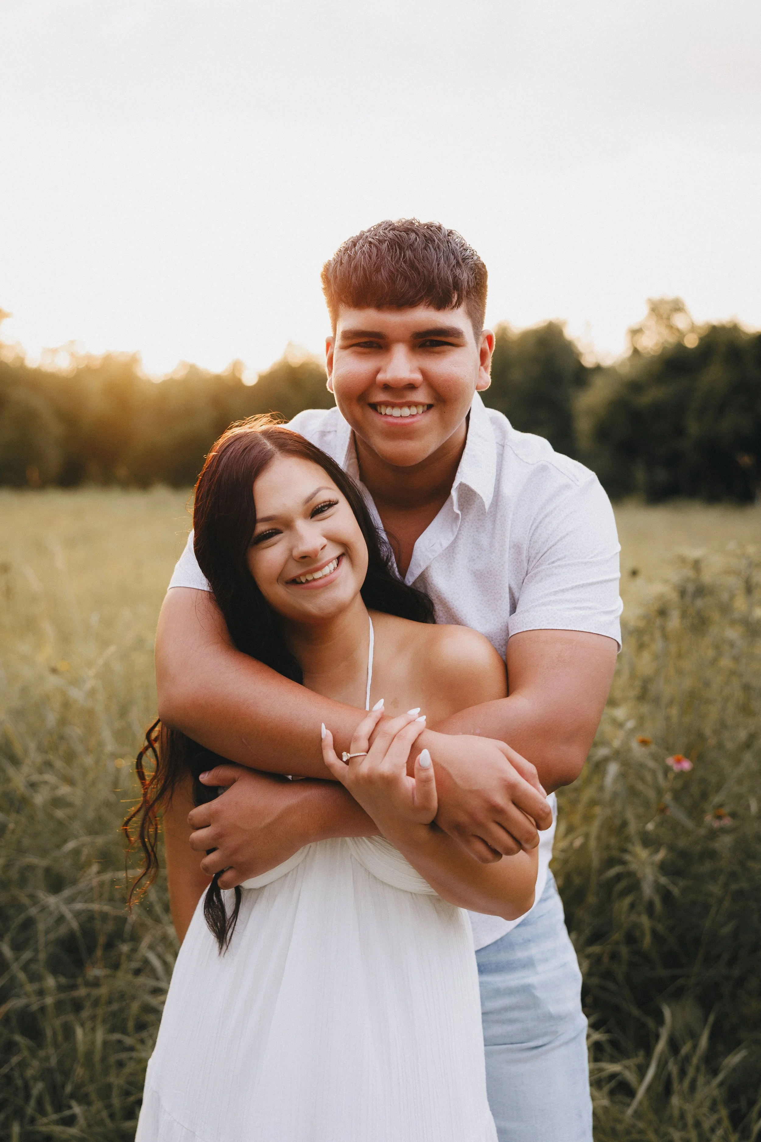 A happy young couple embracing outdoors in a grassy field during sunset, smiling at the camera.  Shot in Topeka, Kansas. 
