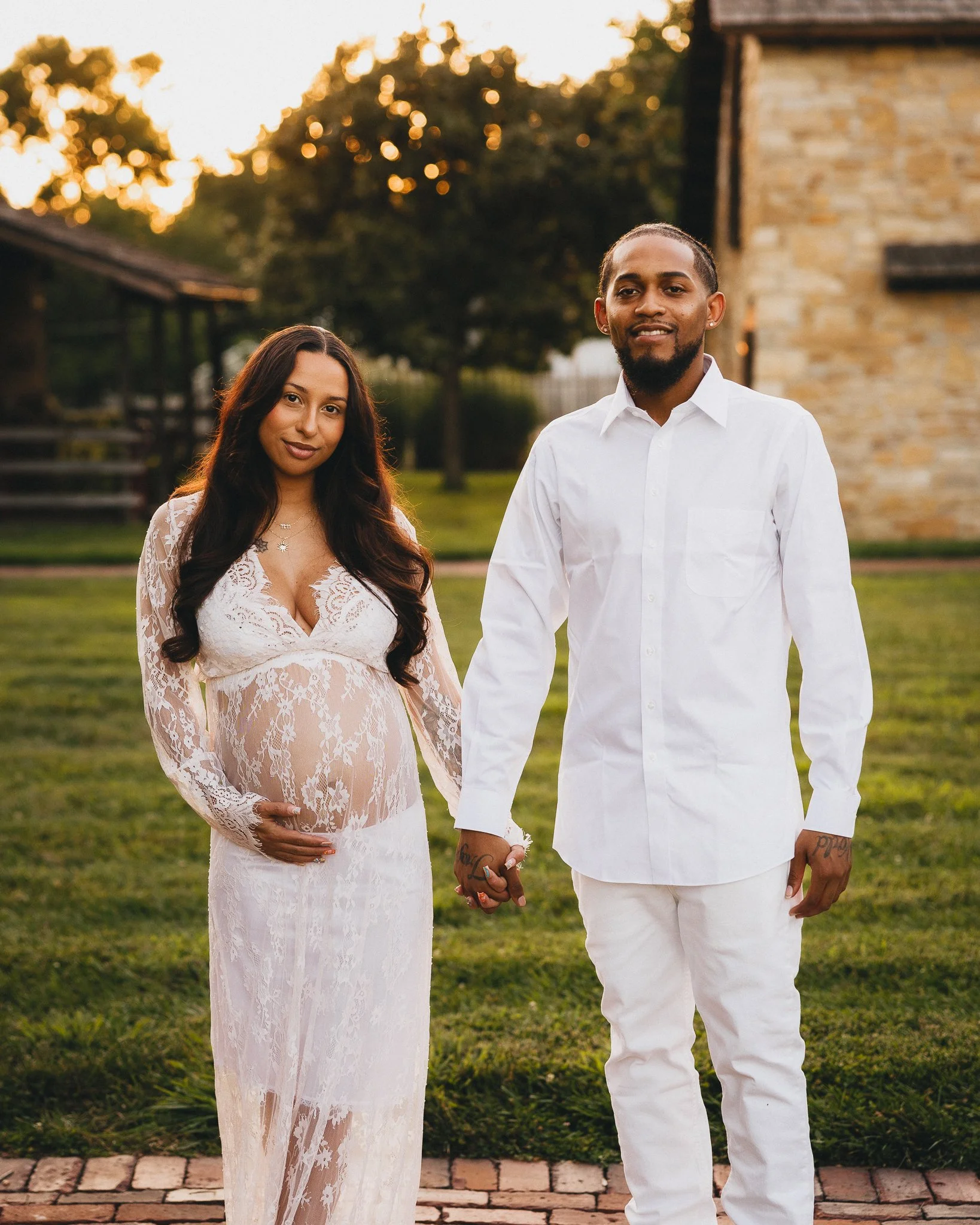 A pregnant woman and a man are standing hand in hand outdoors during sunset. The woman is wearing a sheer, white lace dress, and the man is dressed in a white shirt and white pants. They are holding hands and smiling, with trees and a brick building 