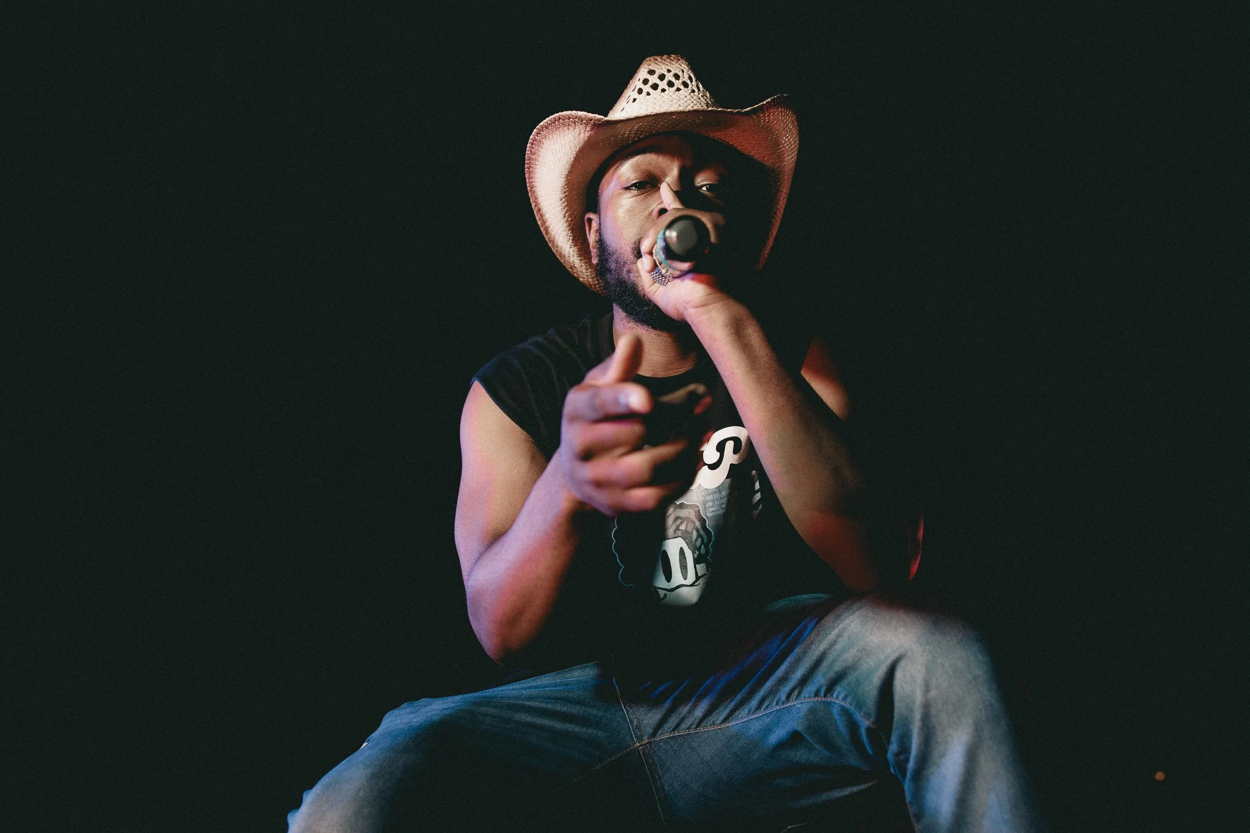 A man wearing a cowboy hat, a sleeveless black shirt, and jeans is sitting and holding a microphone up to his face, pointing towards the camera against a dark background. Shot at Fiesta Topeka.