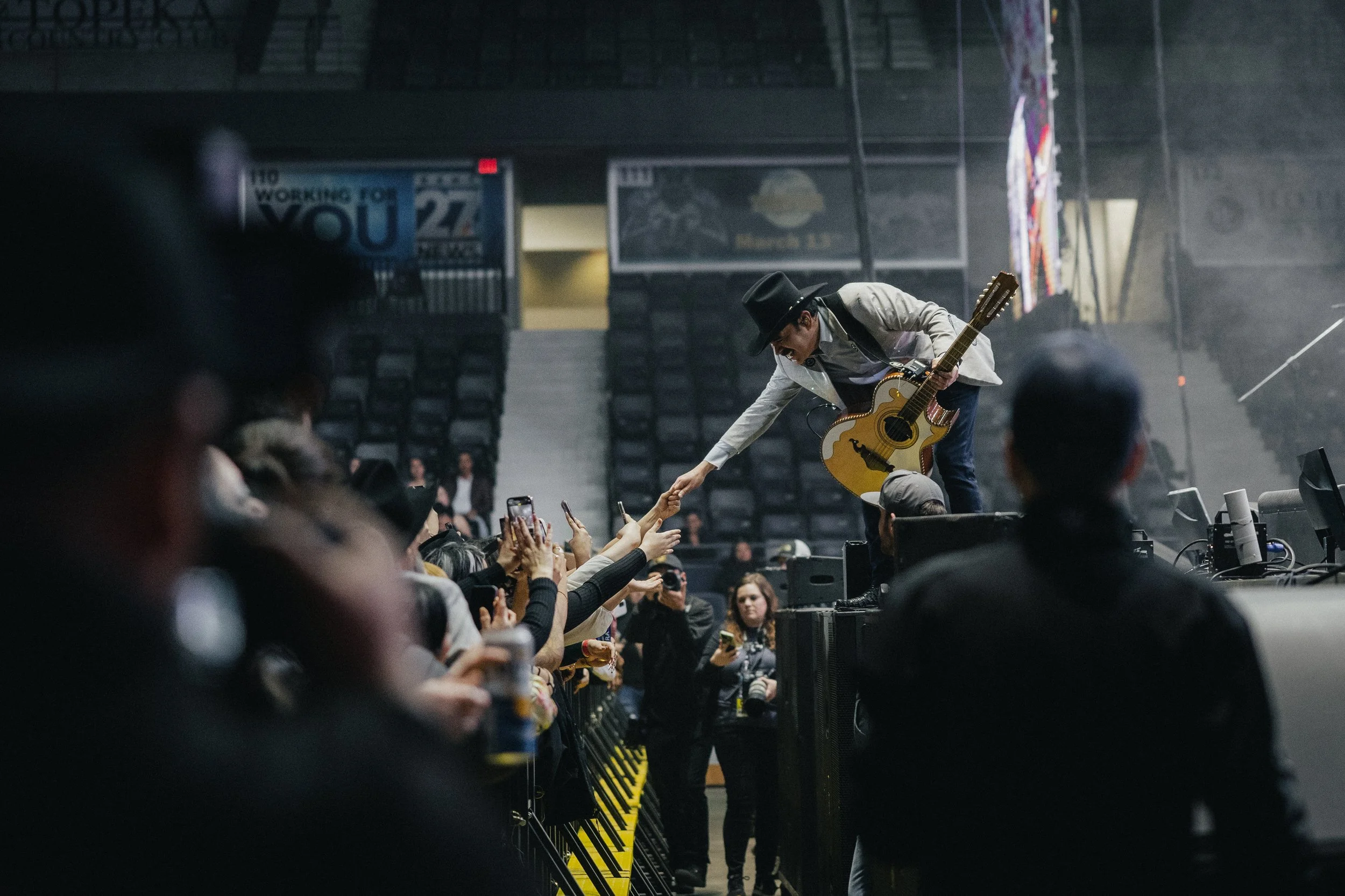 A musician in a gray blazer and black hat leans over a barricade to shake hands with fans during a concert. The musician holds an acoustic guitar, and fans are reaching out with their hands and phones to touch or take photos. The background shows a s