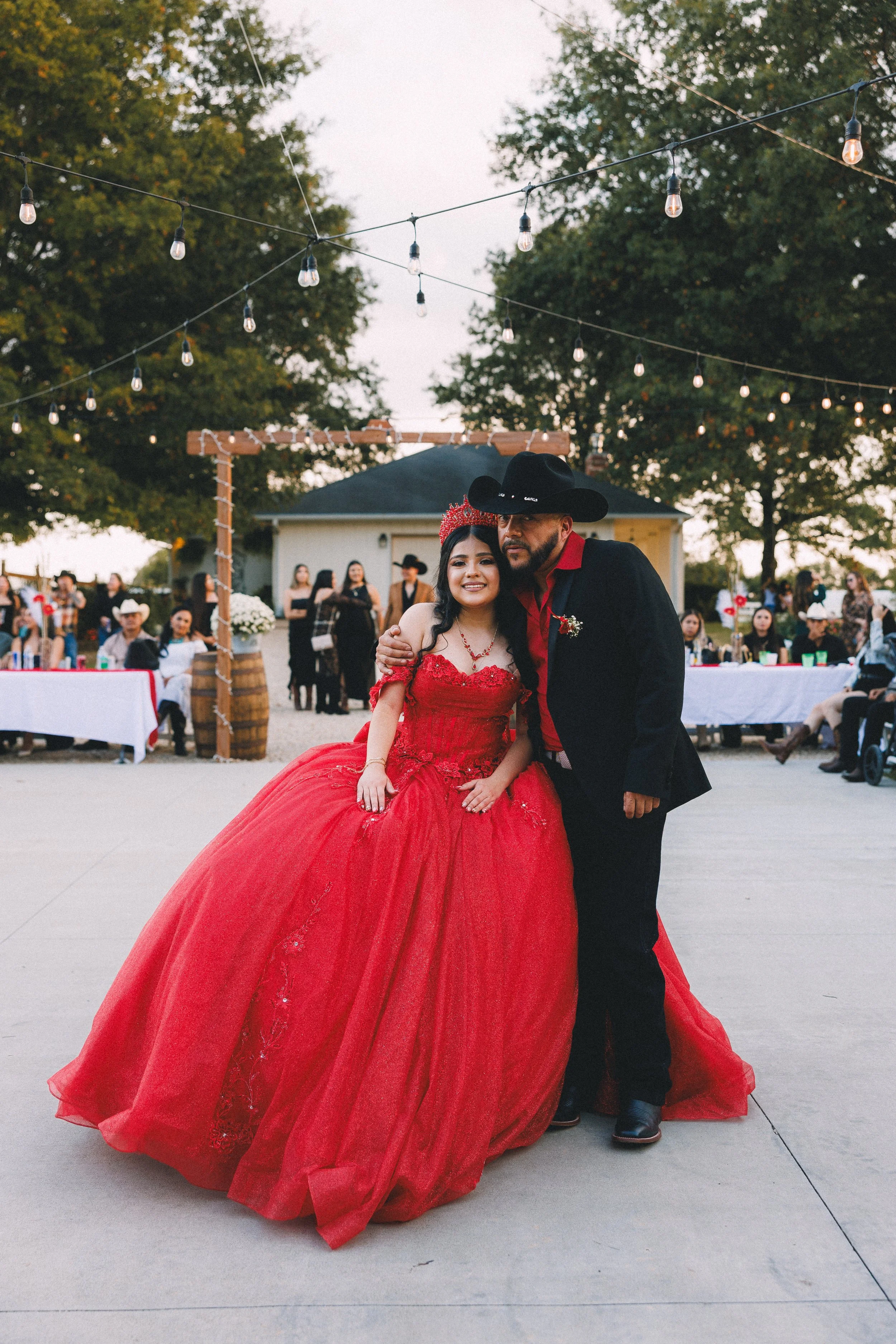 A young woman in a red ball gown and a man in a black cowboy hat and suit pose together at an outdoor event, likely a quinceañera or Sweet 16 celebration, with guests and string lights in the background.
