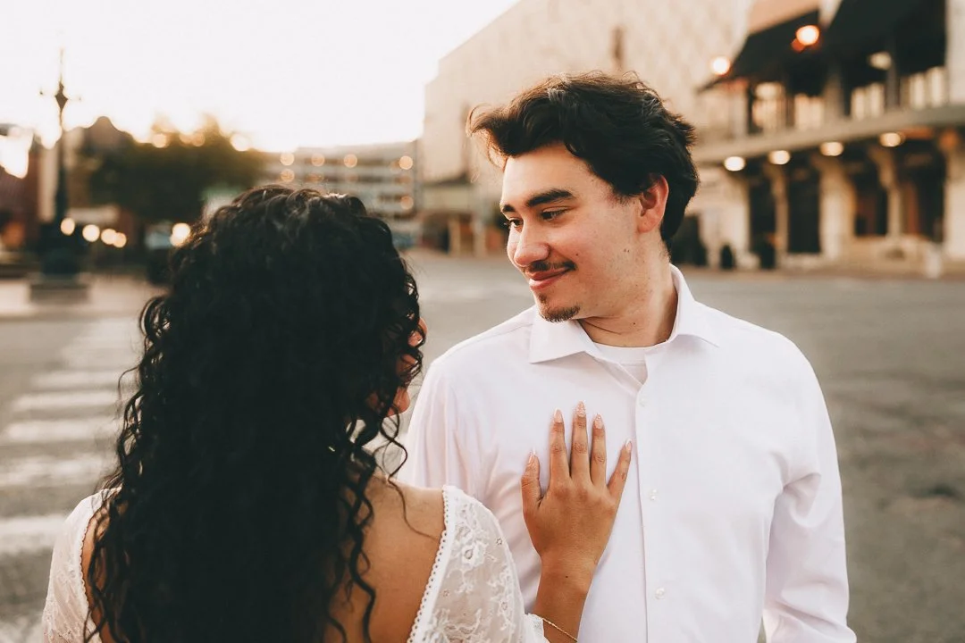A couple standing close together outdoors, with the woman touching the man's chest and both looking at each other, in an urban setting during sunset. Shot at Kansas City, Country Club Plaza.