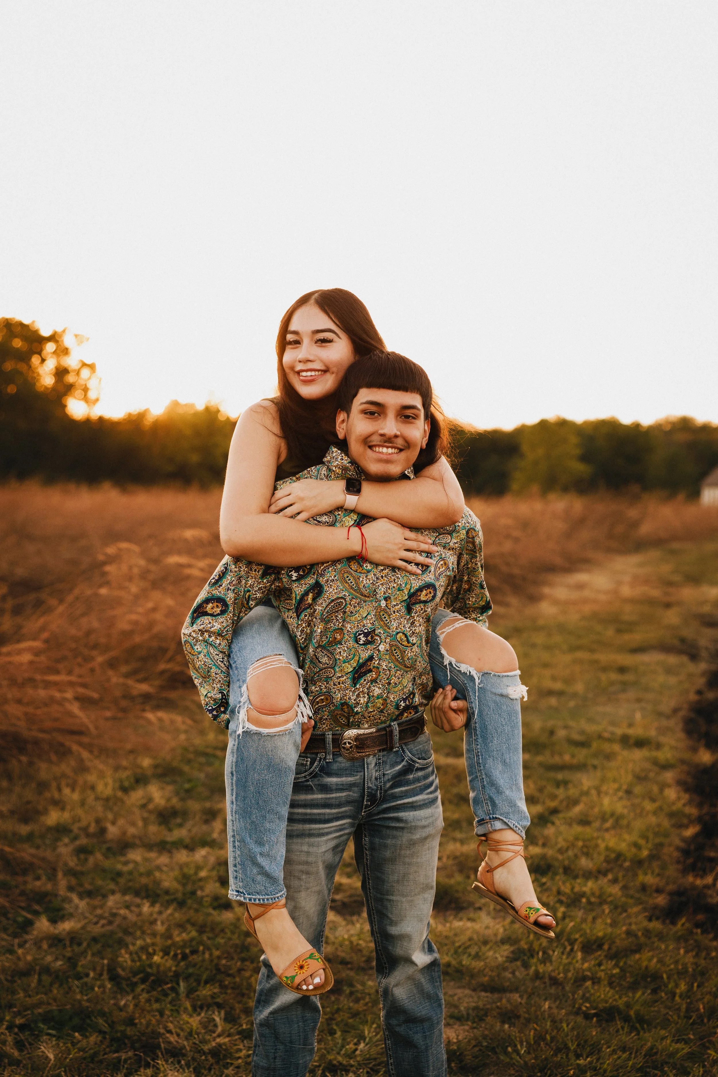 A young couple outdoors in a grassy field during sunset. The man is carrying the woman on his back, both smiling and enjoying the moment. Shot in Topeka, Kansas. 