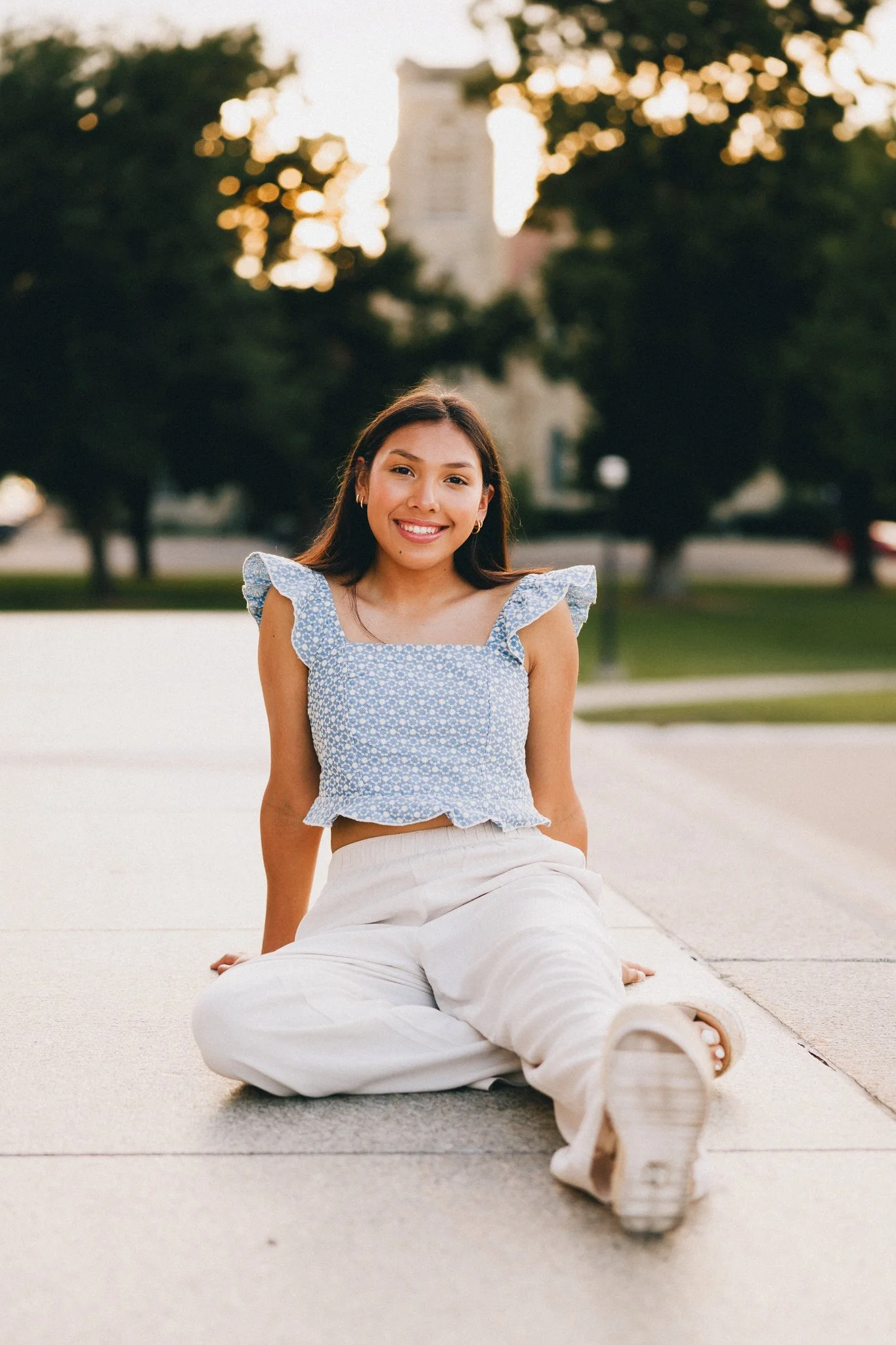 A young woman sitting on the sidewalk outdoors in front of trees and a building at sunset, smiling at the camera.  Shot in Topeka, Kansas. 