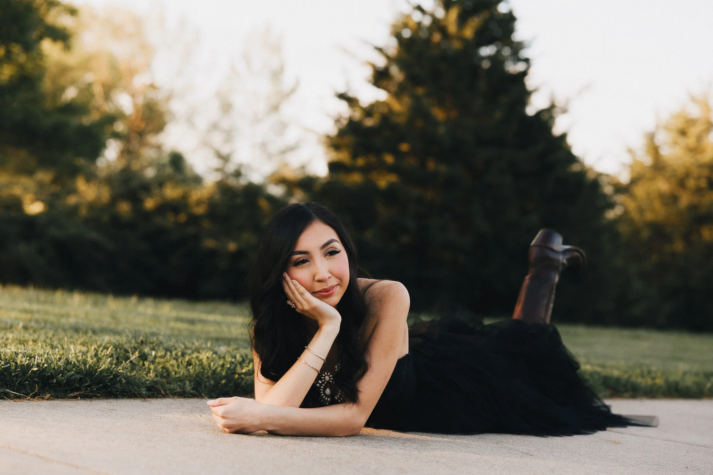A woman with long dark hair lying on her side on a concrete pathway, resting her head on her hand and smiling softly at the camera, outdoors during golden hour with trees in the background.