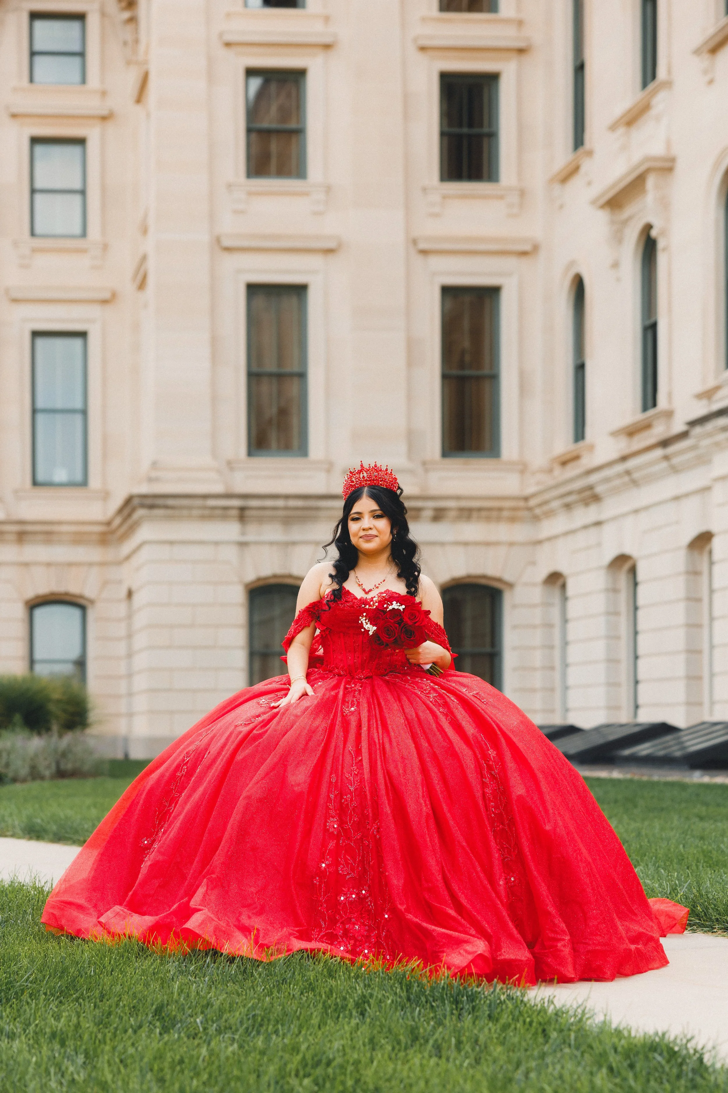 Woman in a red gown holding a bouquet of red roses standing on grass in front of a beige building.