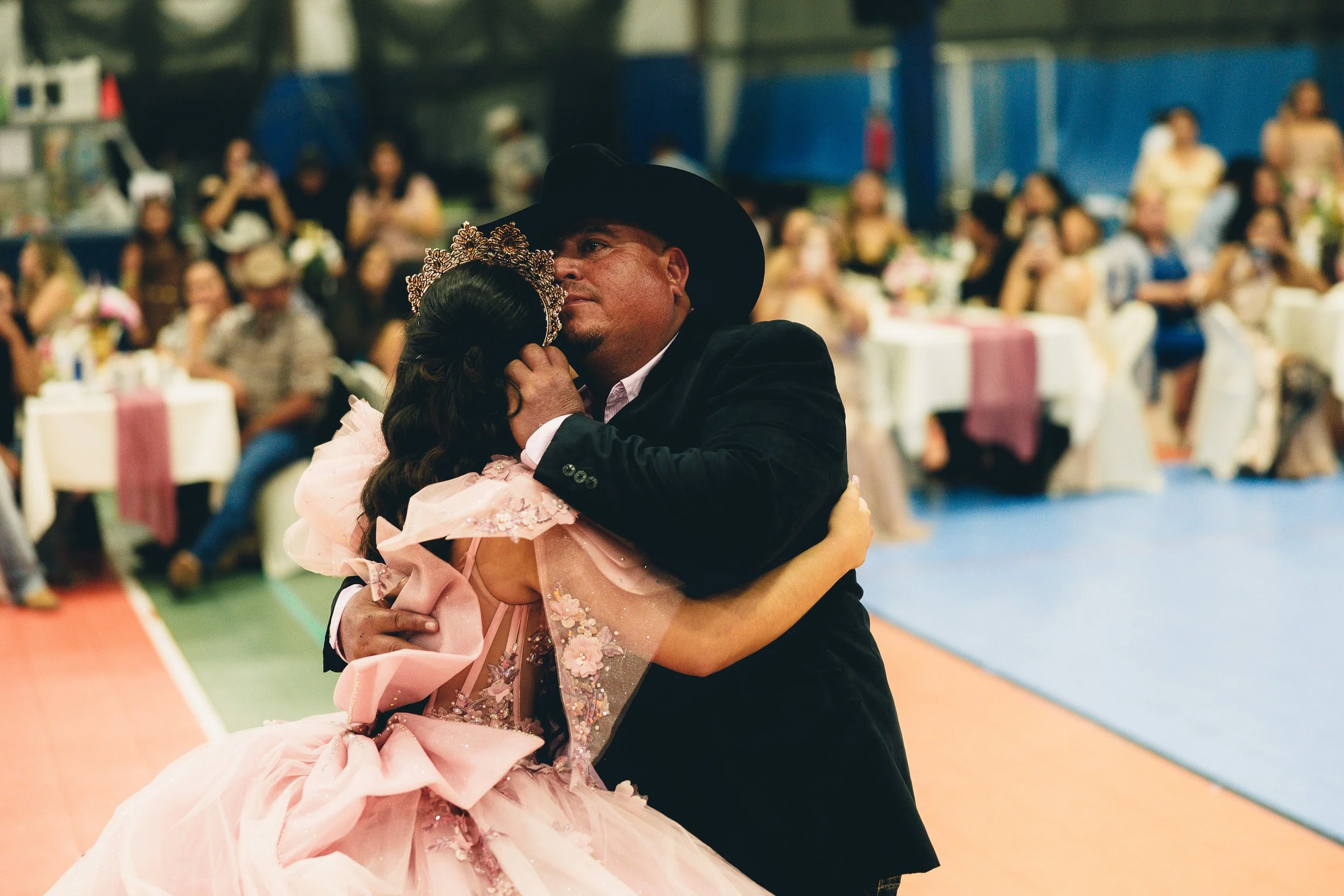 A man and a woman sharing a dance at a celebratory event, with the woman wearing a pink dress and a tiara, and the man wearing a black hat and suit, surrounded by seated guests.