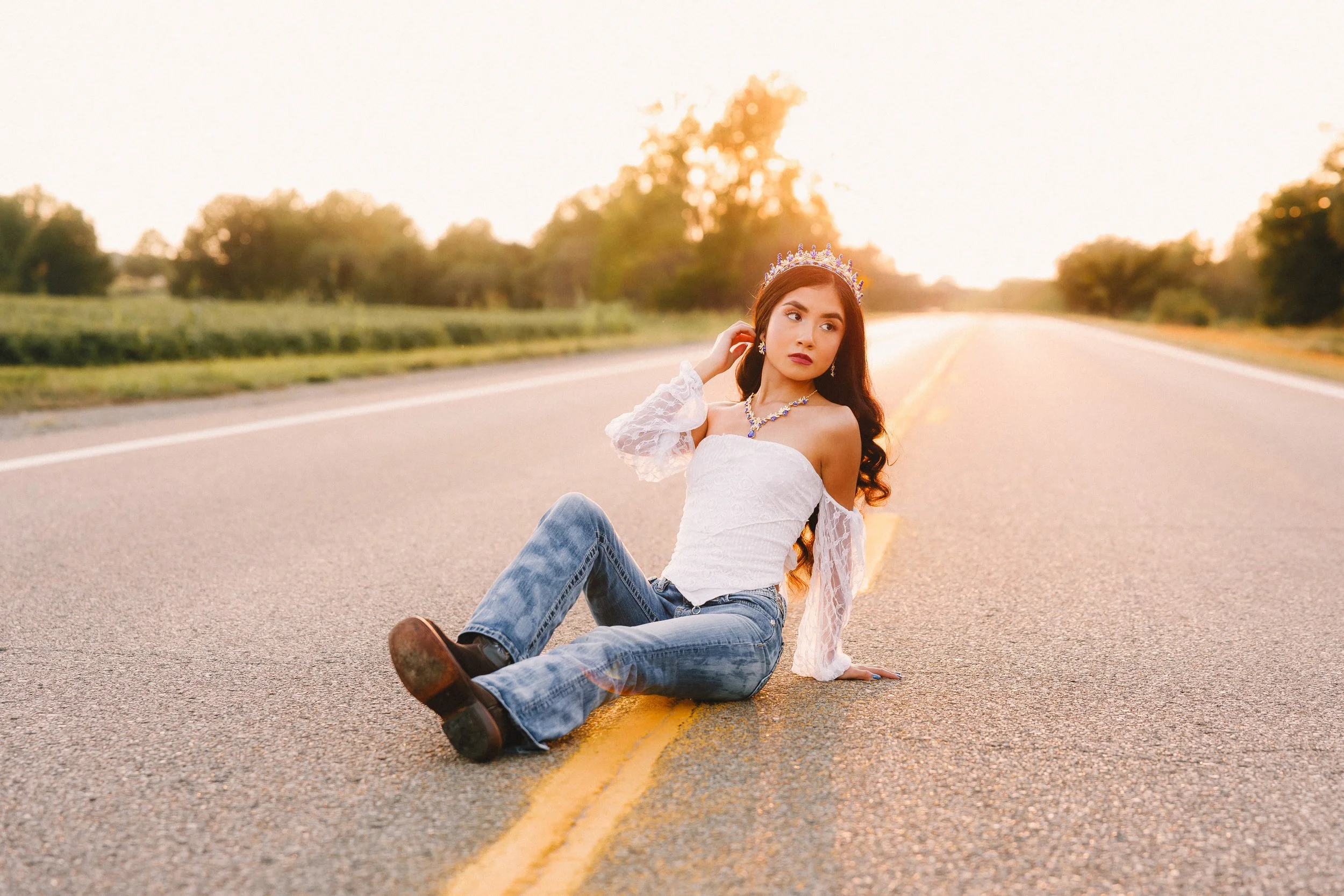 A woman in a white off-shoulder top and jeans sitting in the middle of an empty road at sunset, wearing a crown and jewelry, with trees in the background.