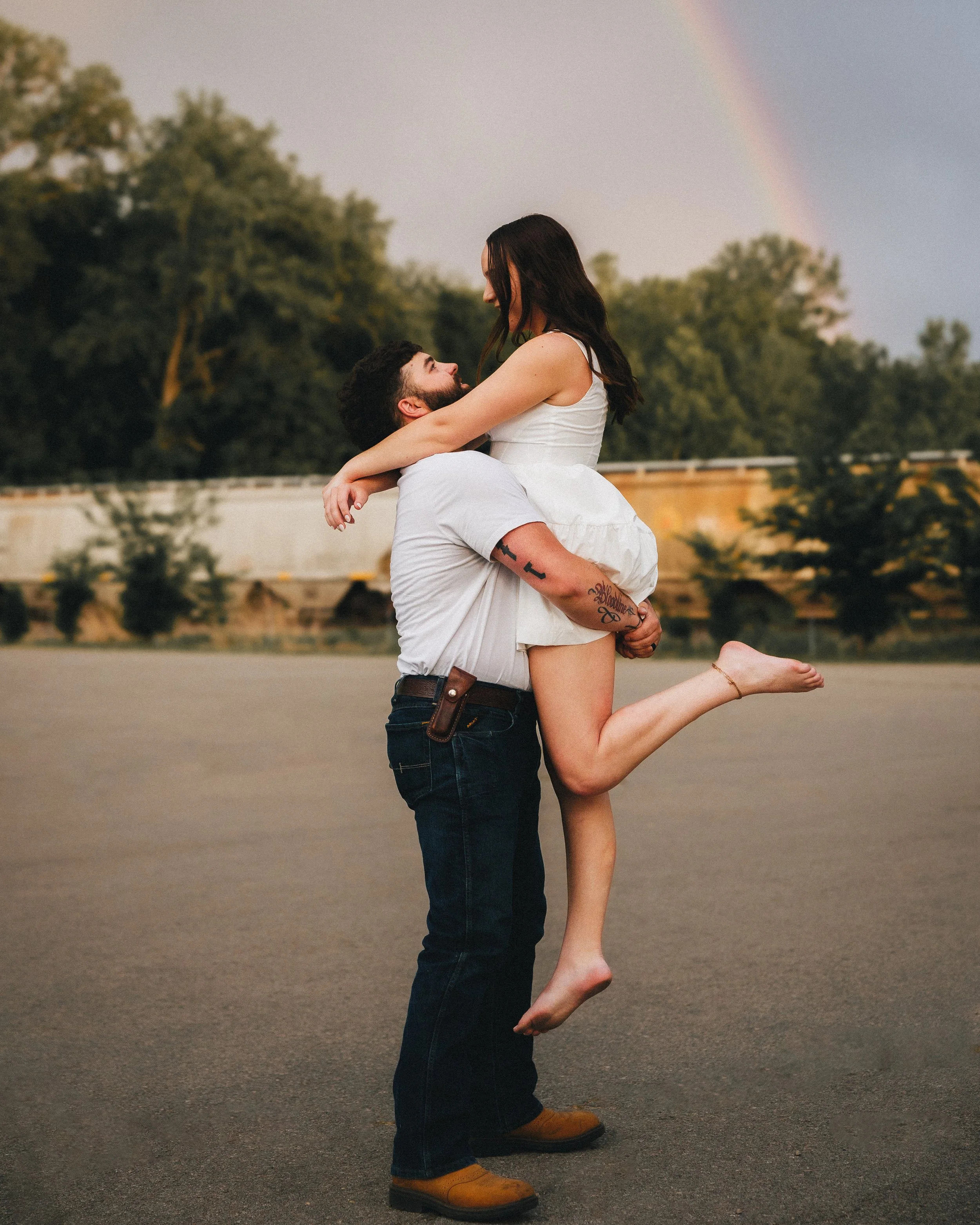 A man holding a woman in his arms outdoors on a road, with a rainbow and trees in the background at dusk.