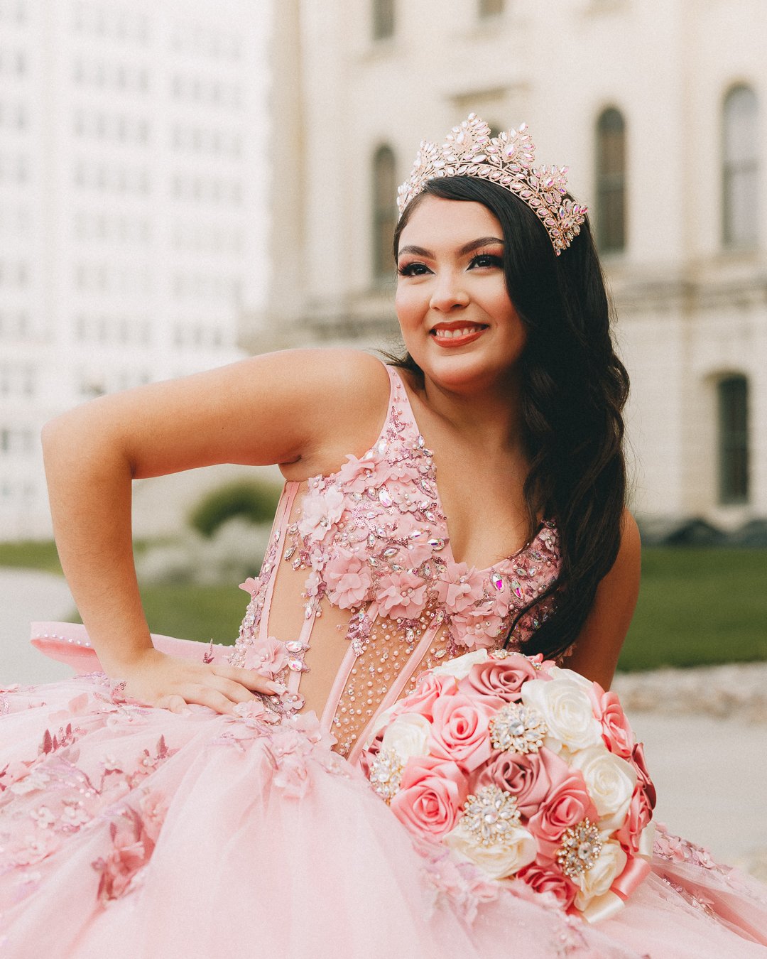 Young woman in a pink, embellished quinceañera dress with a tiara, holding a bouquet of pink and white roses, standing outdoors with a building in the background.