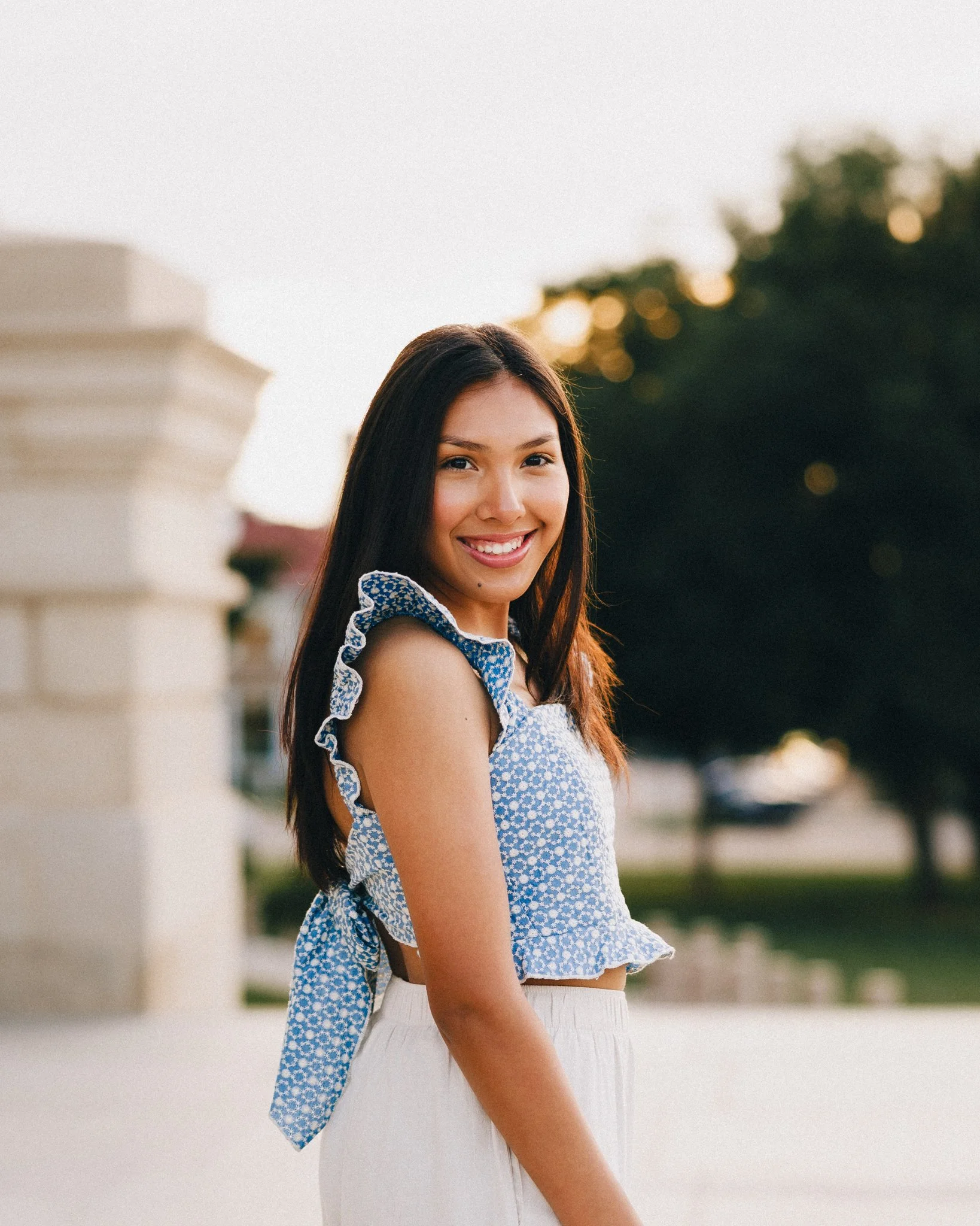 A young woman in a sleeveless blue floral top and white pants smiling outdoors during sunset.  Shot in Topeka, Kansas. 