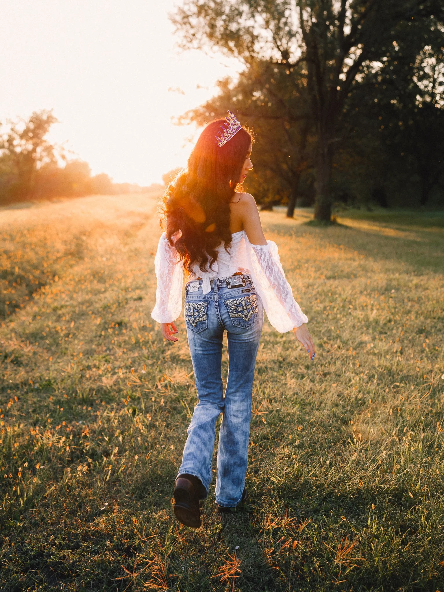A young woman walking away in a grassy field during sunset, wearing a crown, off-shoulder white top, and blue jeans.