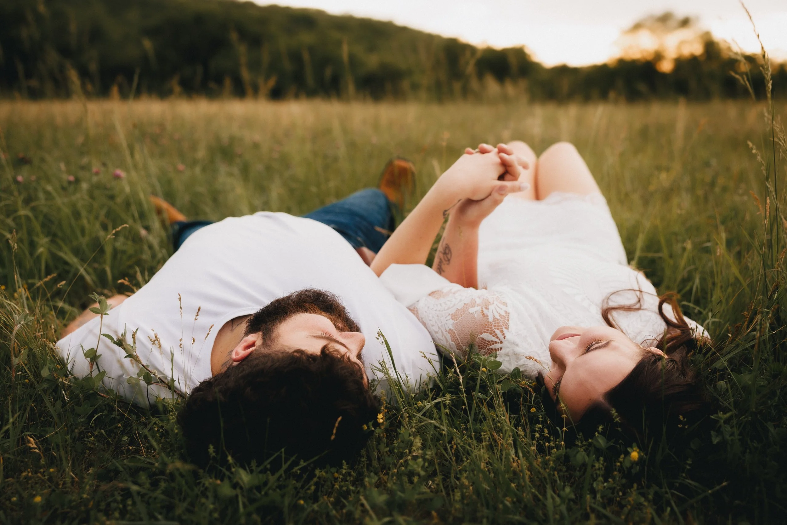 A couple lying on their backs in a grassy field holding hands and looking at each other with closed eyes during sunset.