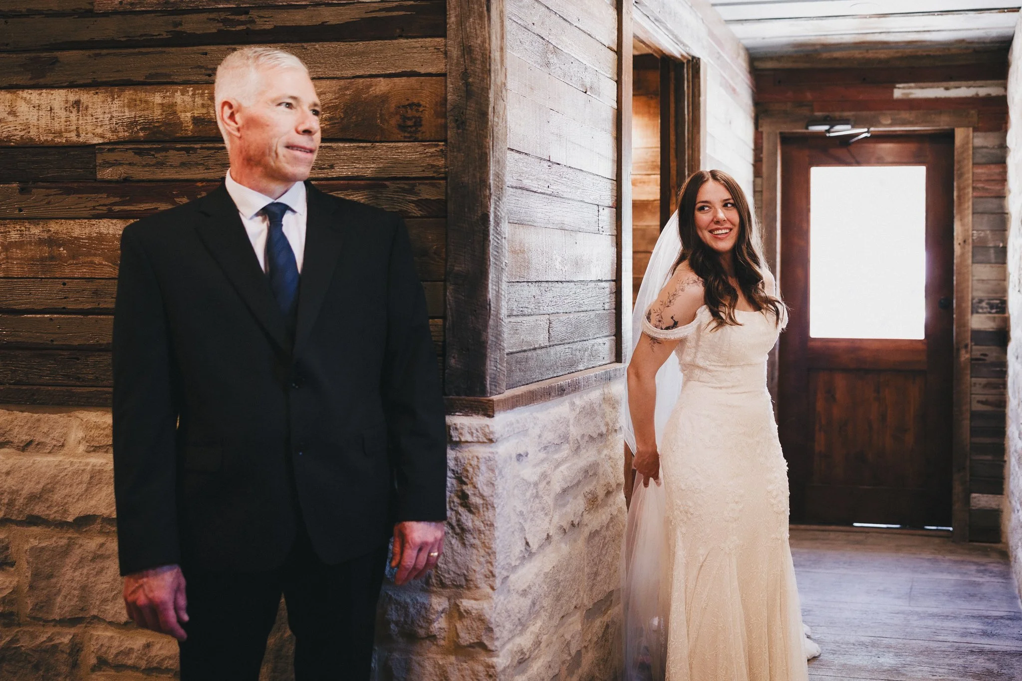 Wedding scene with an older man in a black suit, white shirt, and navy tie, standing to the left, and a young woman in a cream lace wedding dress with off-shoulder sleeves, holding a veil, smiling at the camera, in a rustic wooden interior.