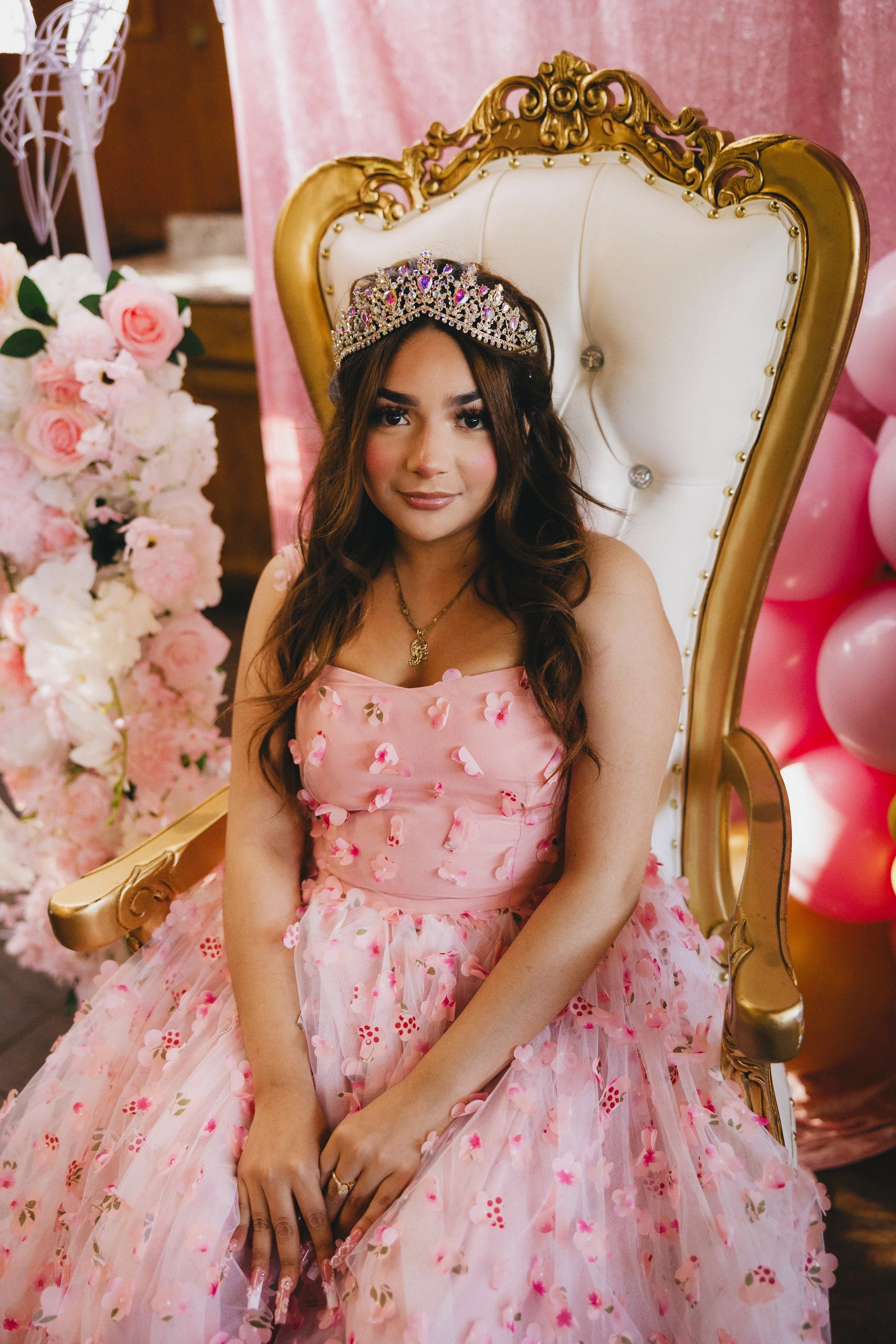 A young woman wearing a pink dress with flower petals, a crown on her head, and sitting on a white and gold throne, celebrating an event with pink and white balloons and flower arrangements.