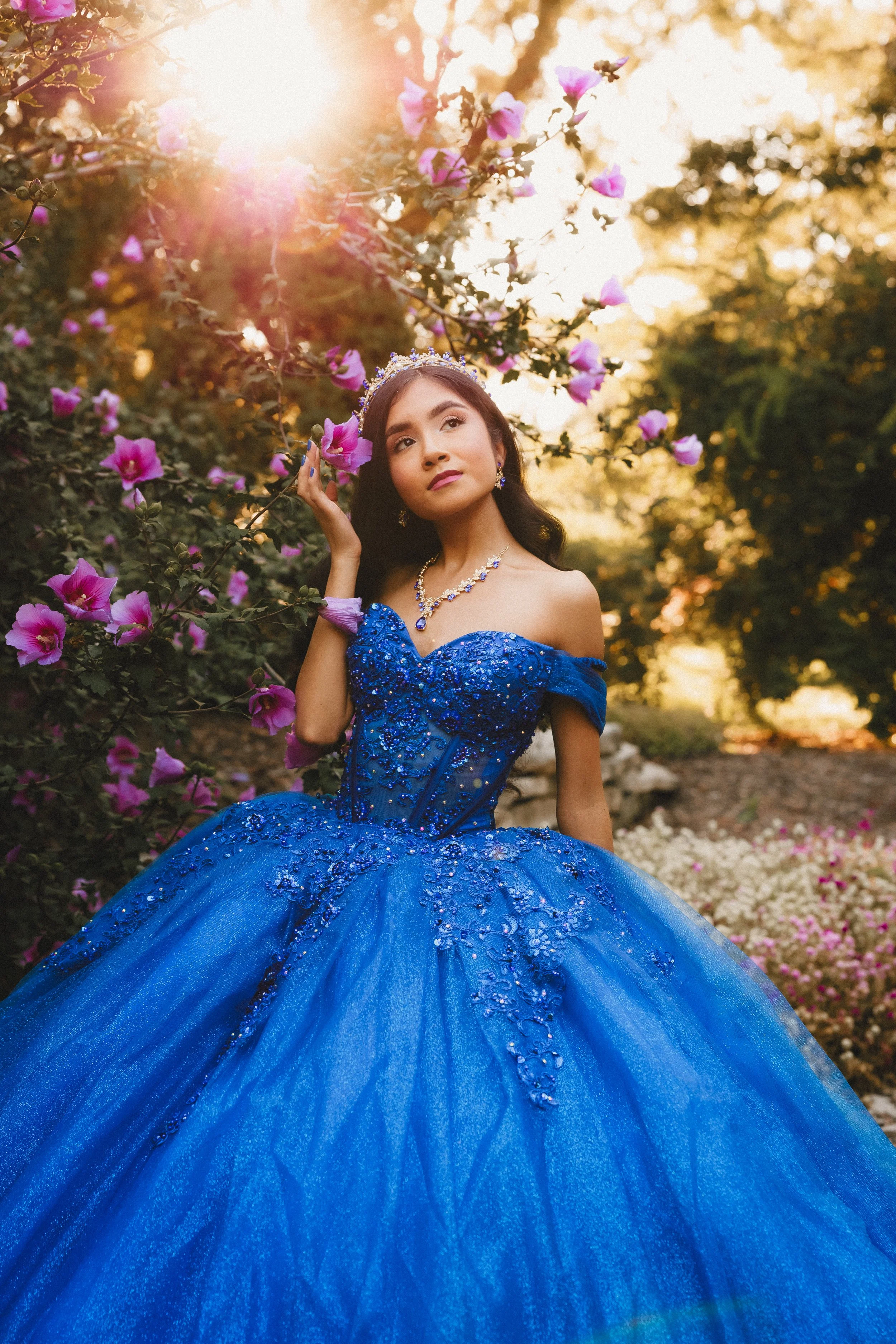 Young quinceañera in a royal blue, embellished dress standing among pink and purple flowers outdoors during sunset.