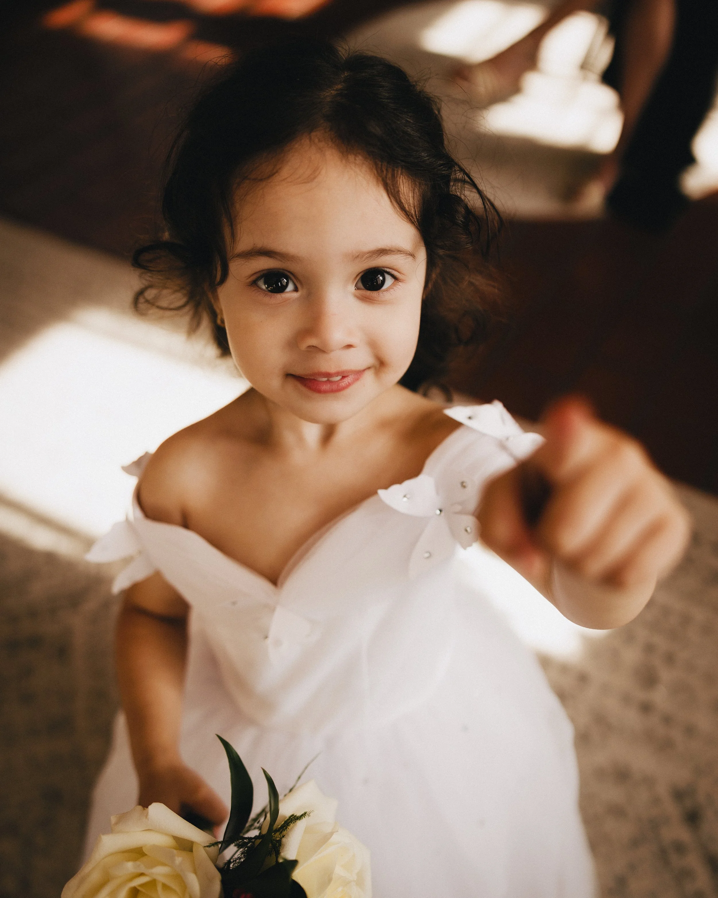 A young girl with dark curly hair and big eyes, wearing a white dress with floral embellishments, holding a bouquet of white flowers, and pointing towards the camera with a smile. Shot at Topeka Kansas venue The Beacon.