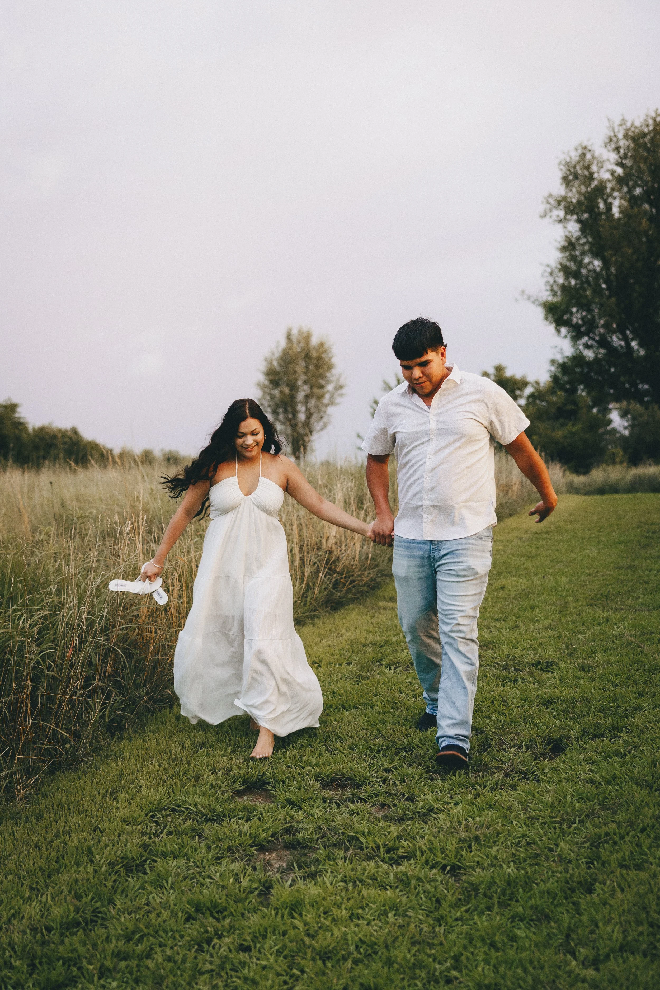 A young couple is walking hand in hand through a grassy field, with the woman smiling and holding her shoes in her left hand.  Shot in Topeka, Kansas. 