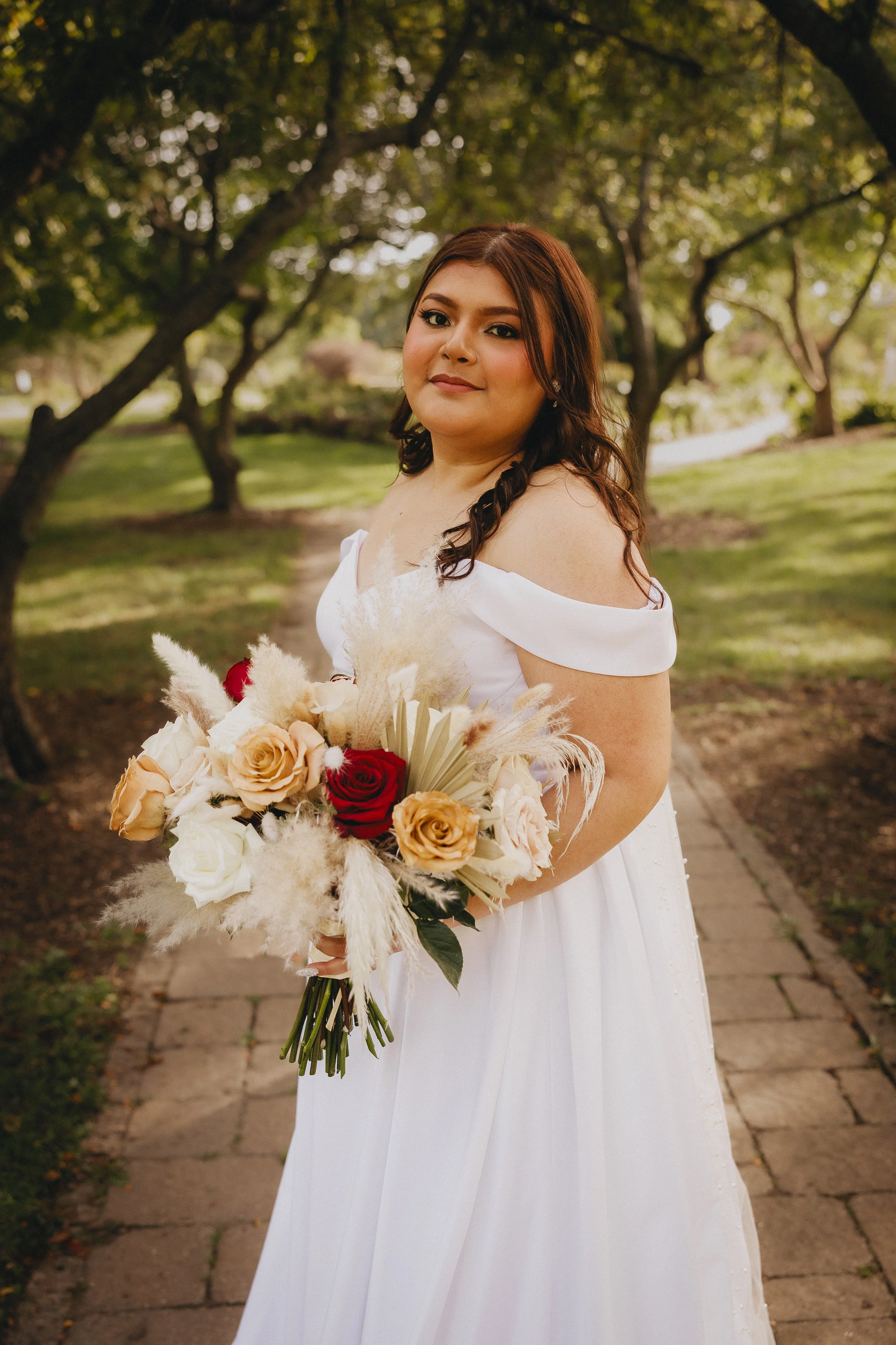 A woman in a white off-shoulder dress holding a bouquet of roses and dried flowers, standing on a brick pathway in a park with trees and green grass.