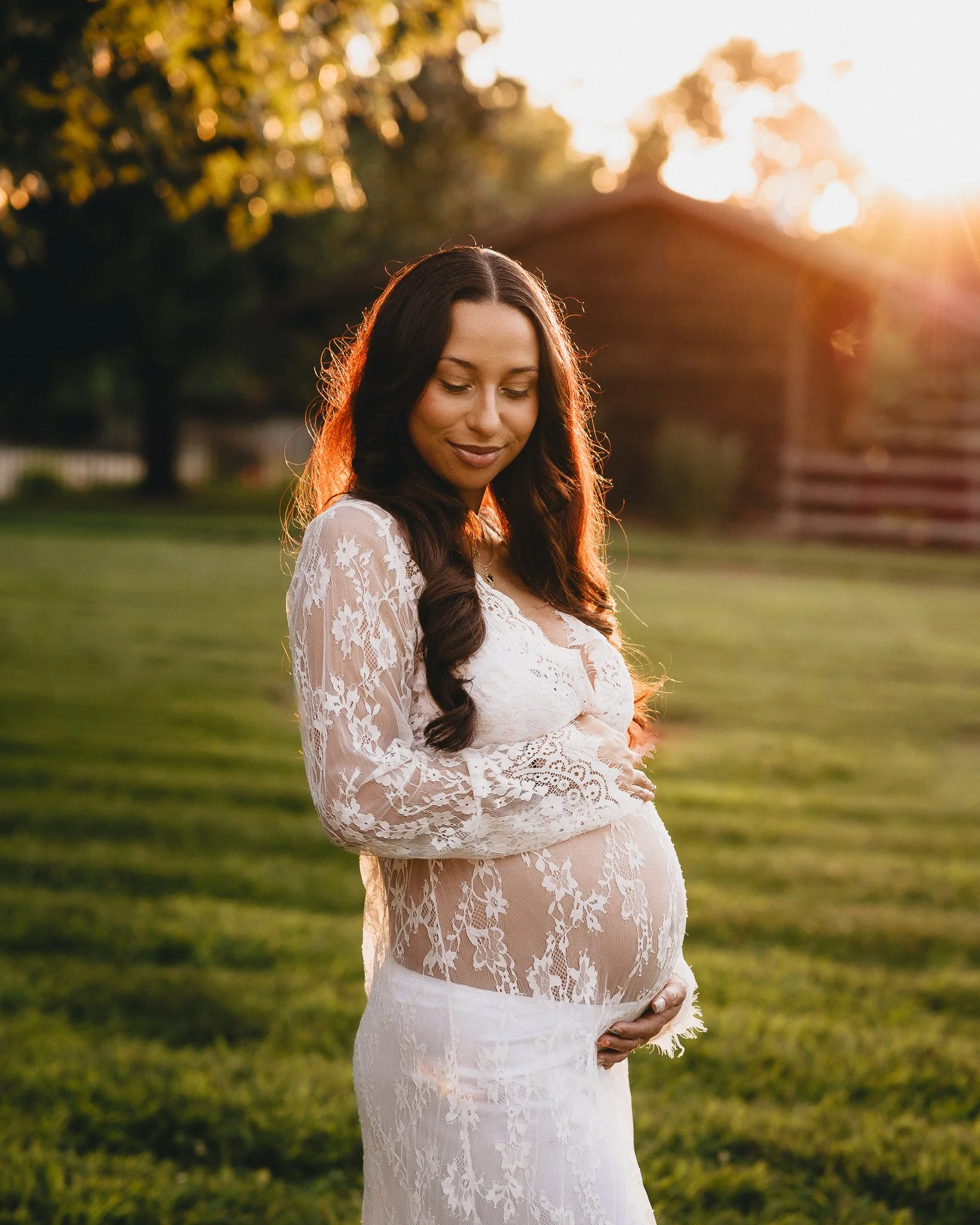 A pregnant woman in a white lace dress standing in a grassy field during sunset, gently holding her belly and smiling with her eyes closed. Shot in Topeka, Kansas. 