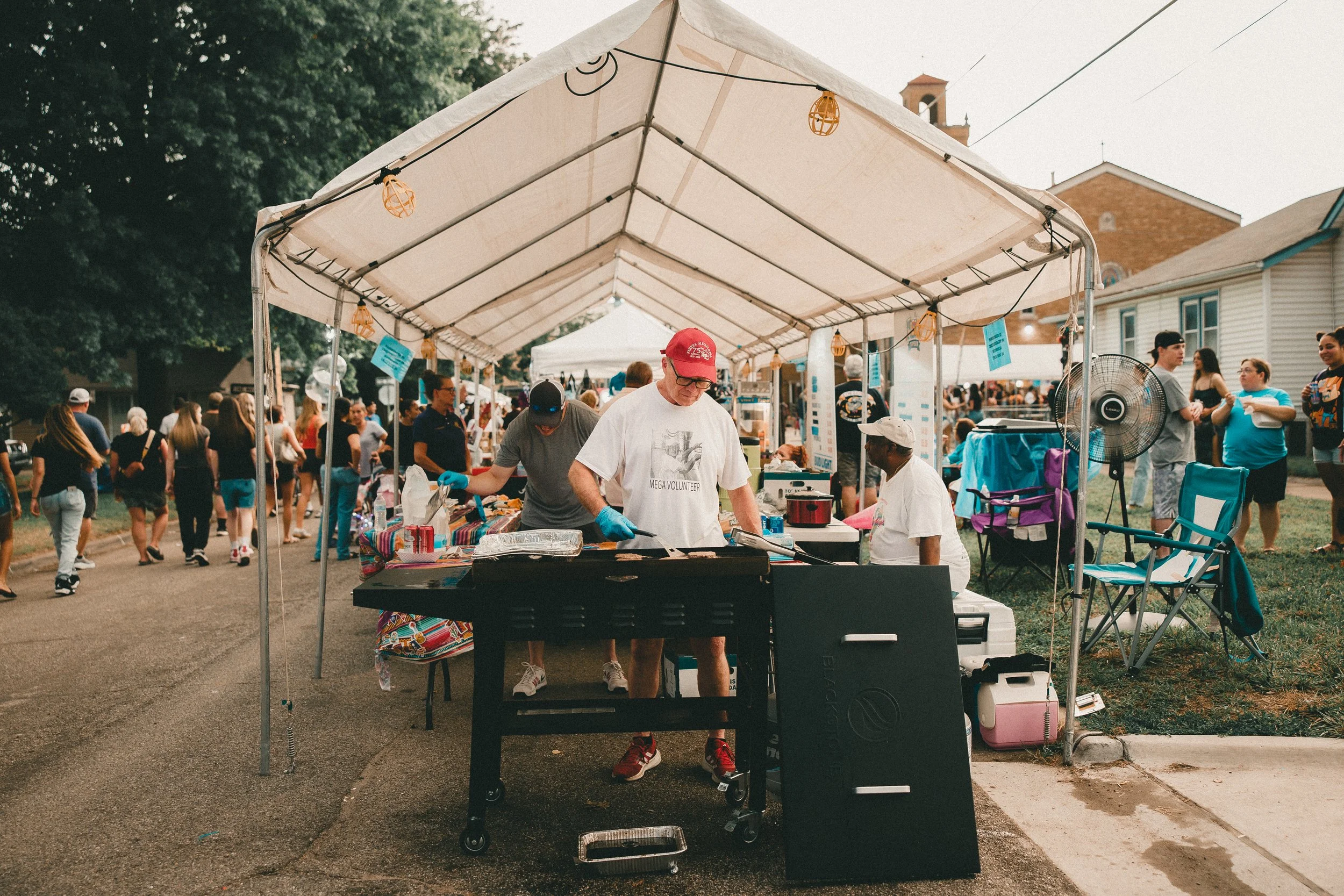 People gathered at an outdoor event with a food stall in the foreground, under a white canopy, with a man cooking on a grill. Several individuals are walking and chatting, and there are houses and trees in the background. Shot at Fiesta Topeka.