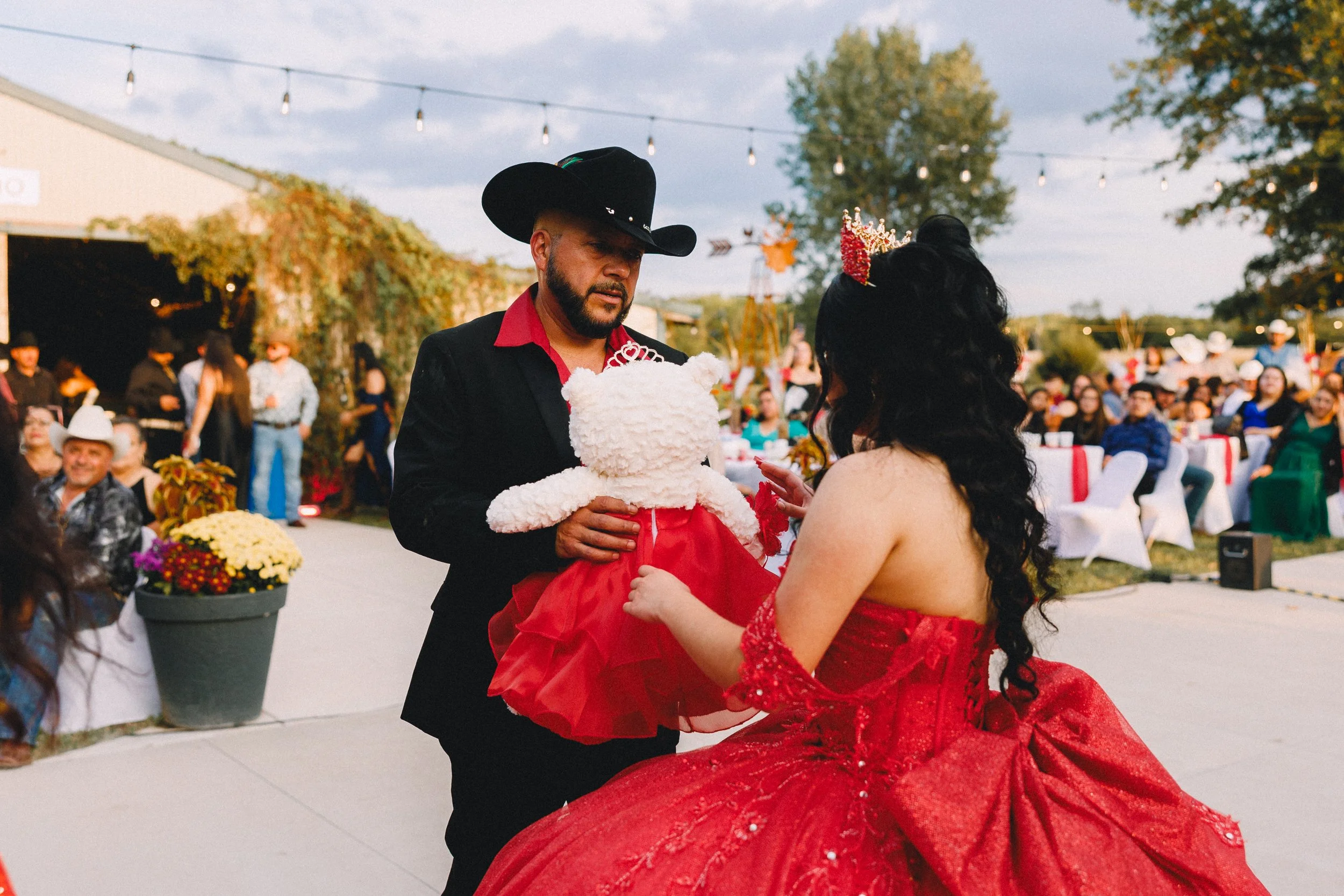 A man in a black cowboy hat and black suit holding a white teddy bear dressed in a red dress. A woman in a red gown with black hair and a red crown is in front of him, with people seated at tables in the background, outdoors during the evening.