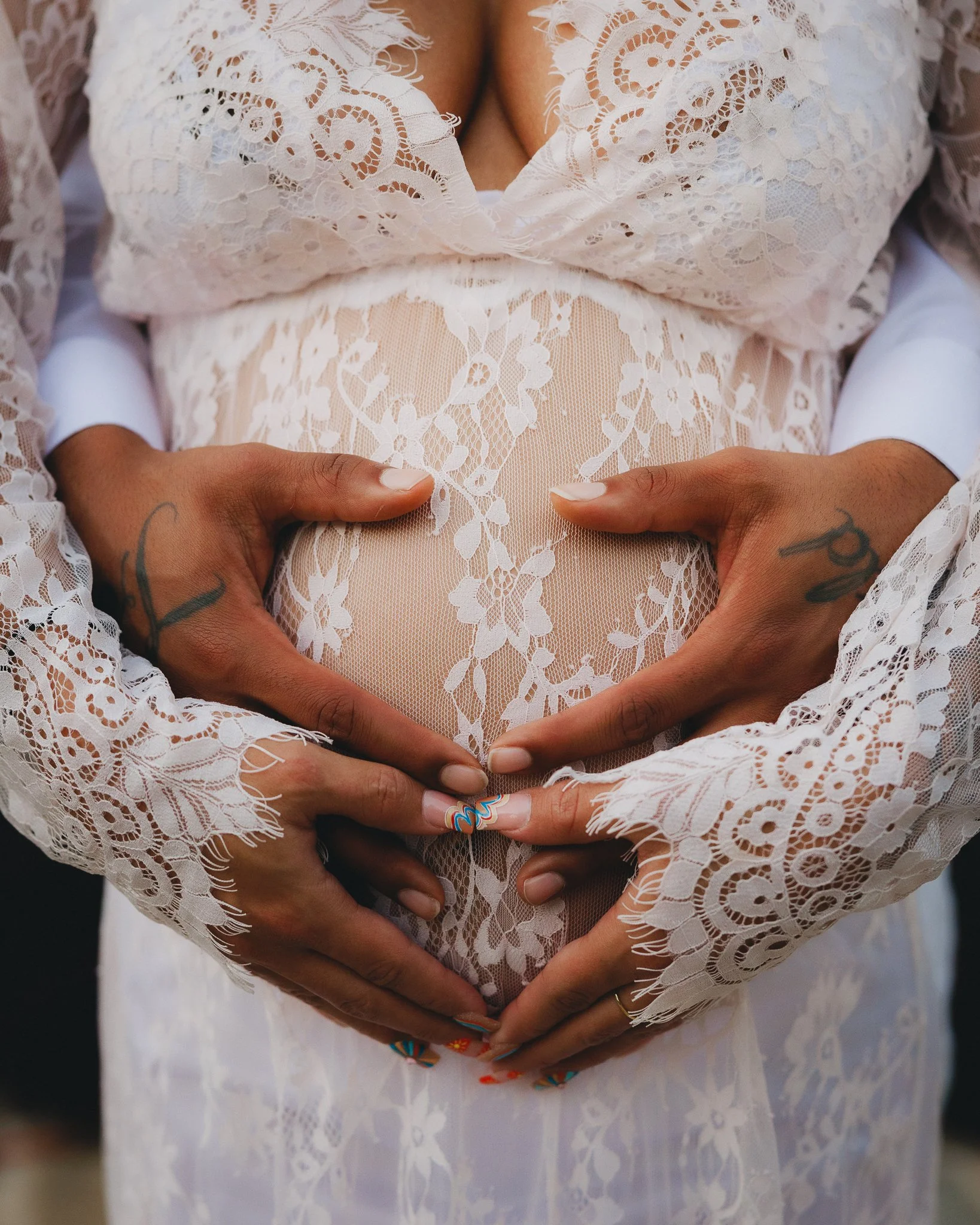 A woman wearing a sheer floral lace dress with her hands forming a heart shape around her pregnant belly. Shot in Topeka, Kansas. 