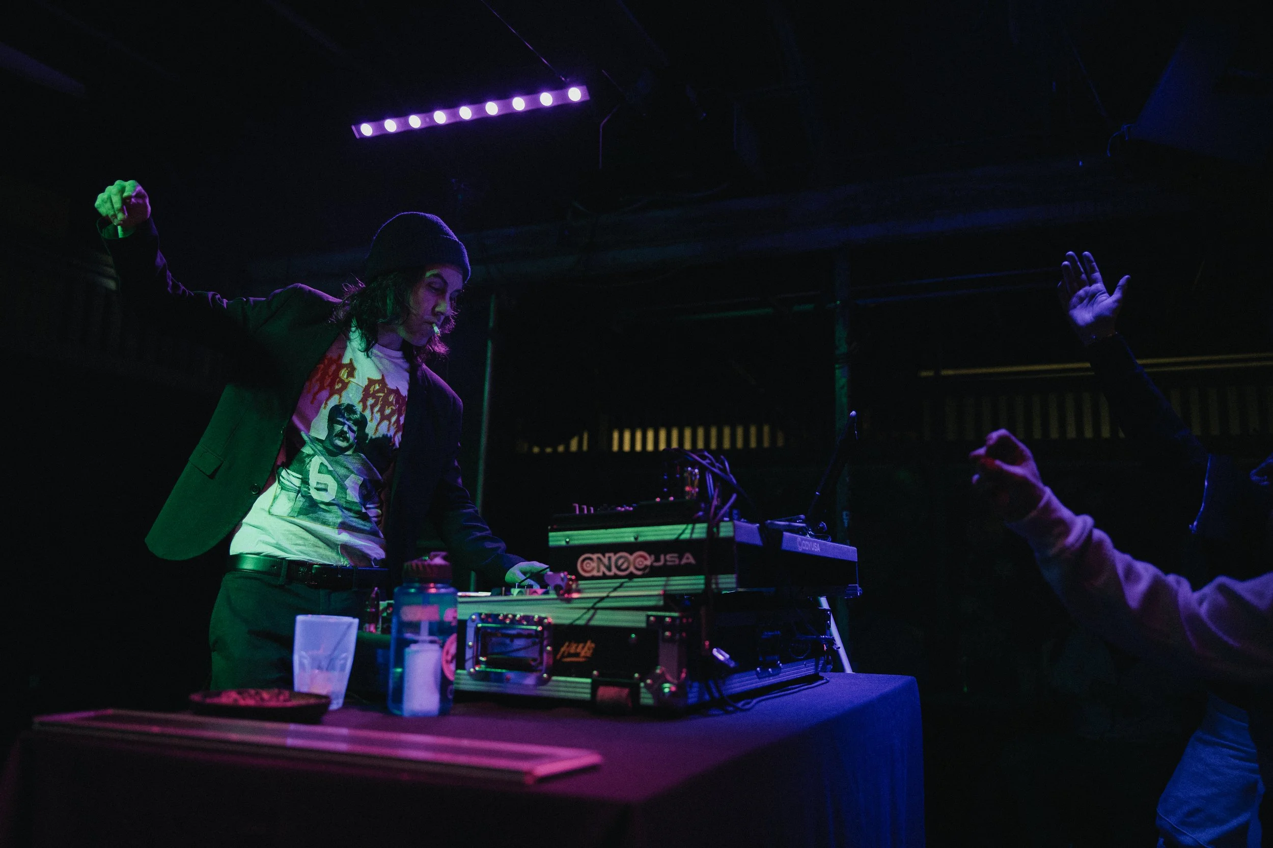 Person in a black jacket and beanie DJing at a table with music equipment, with purple lighting, and another person raising their hands in a dark indoor setting. Shot at The Replay Lounge in Lawrence, Kansas. 