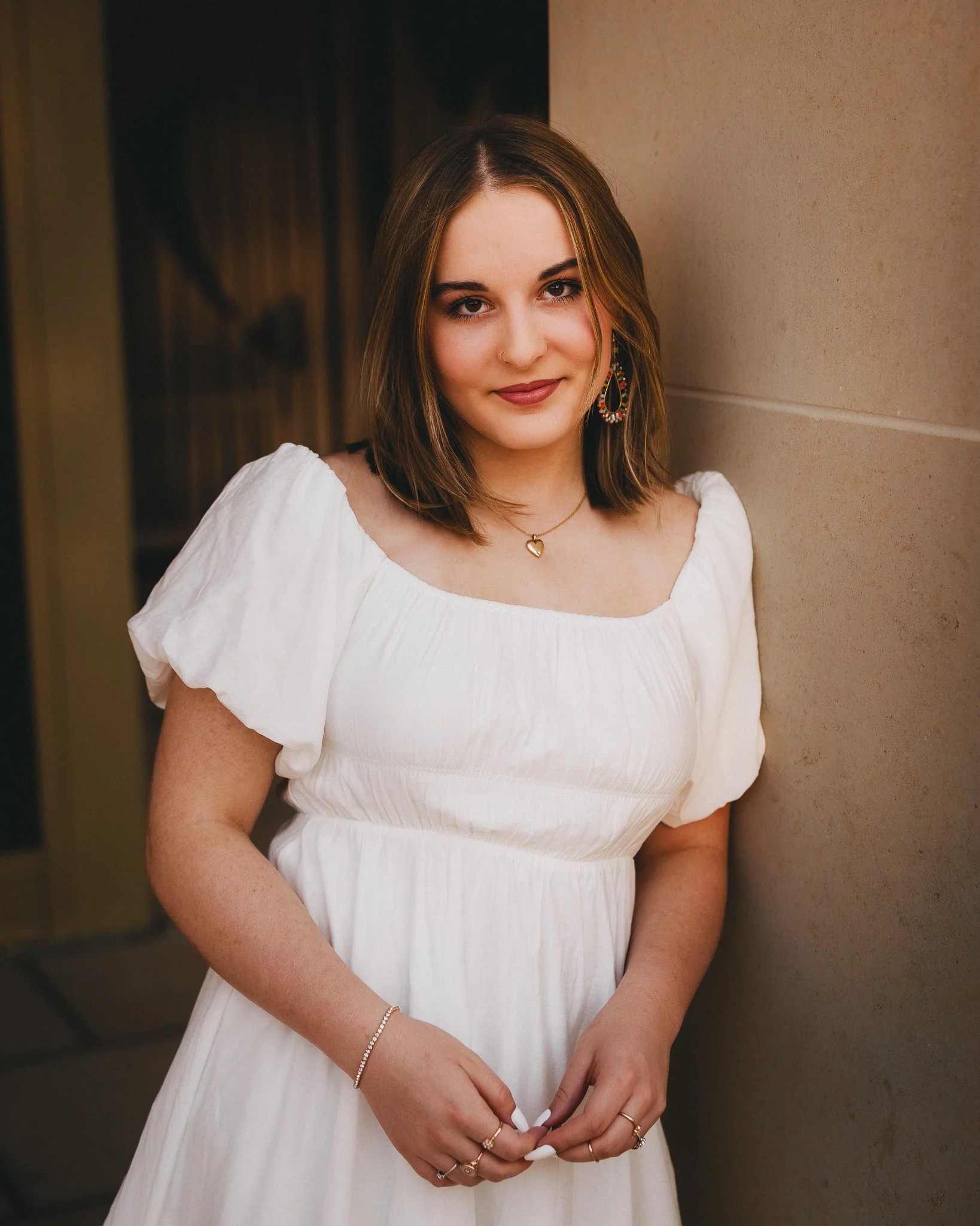 A young woman with shoulder-length brown hair styled in a bob, wearing a white dress with puff sleeves, gold jewelry including earrings, a necklace, and rings, standing against a beige wall, looking at the camera with a slight smile. Shot in Topeka, 