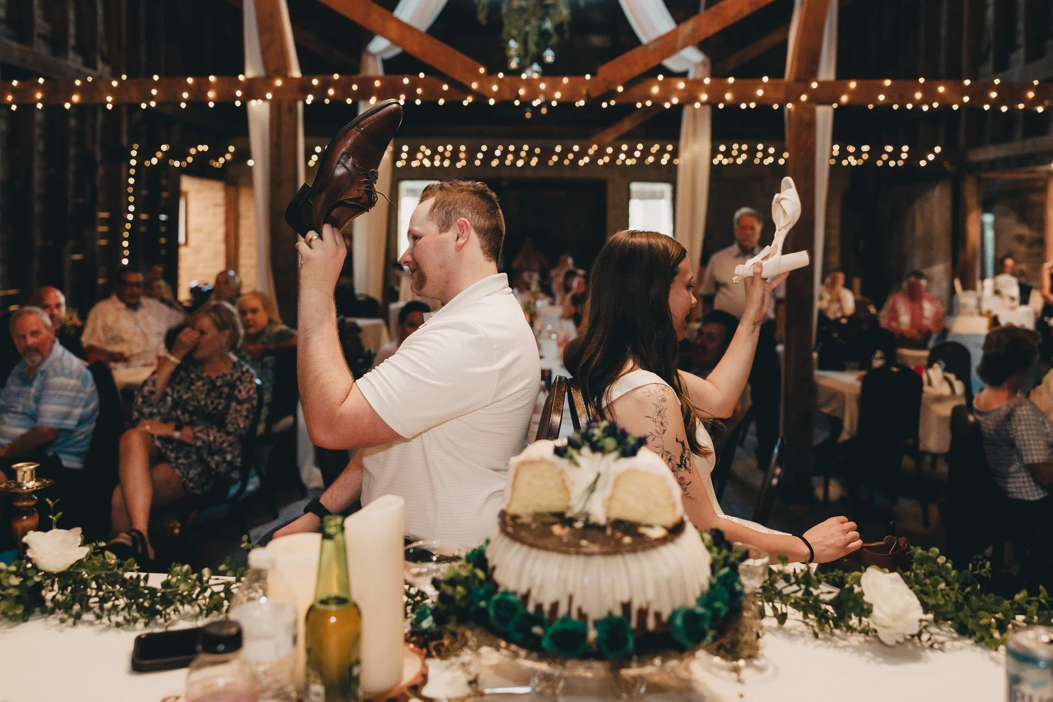 Two people back-to-back, one man and one woman, at a wedding reception, each holding a shoe in one hand and a rolled-up cloth in the other, with guests seated at tables in the background, and a wedding cake in the foreground.