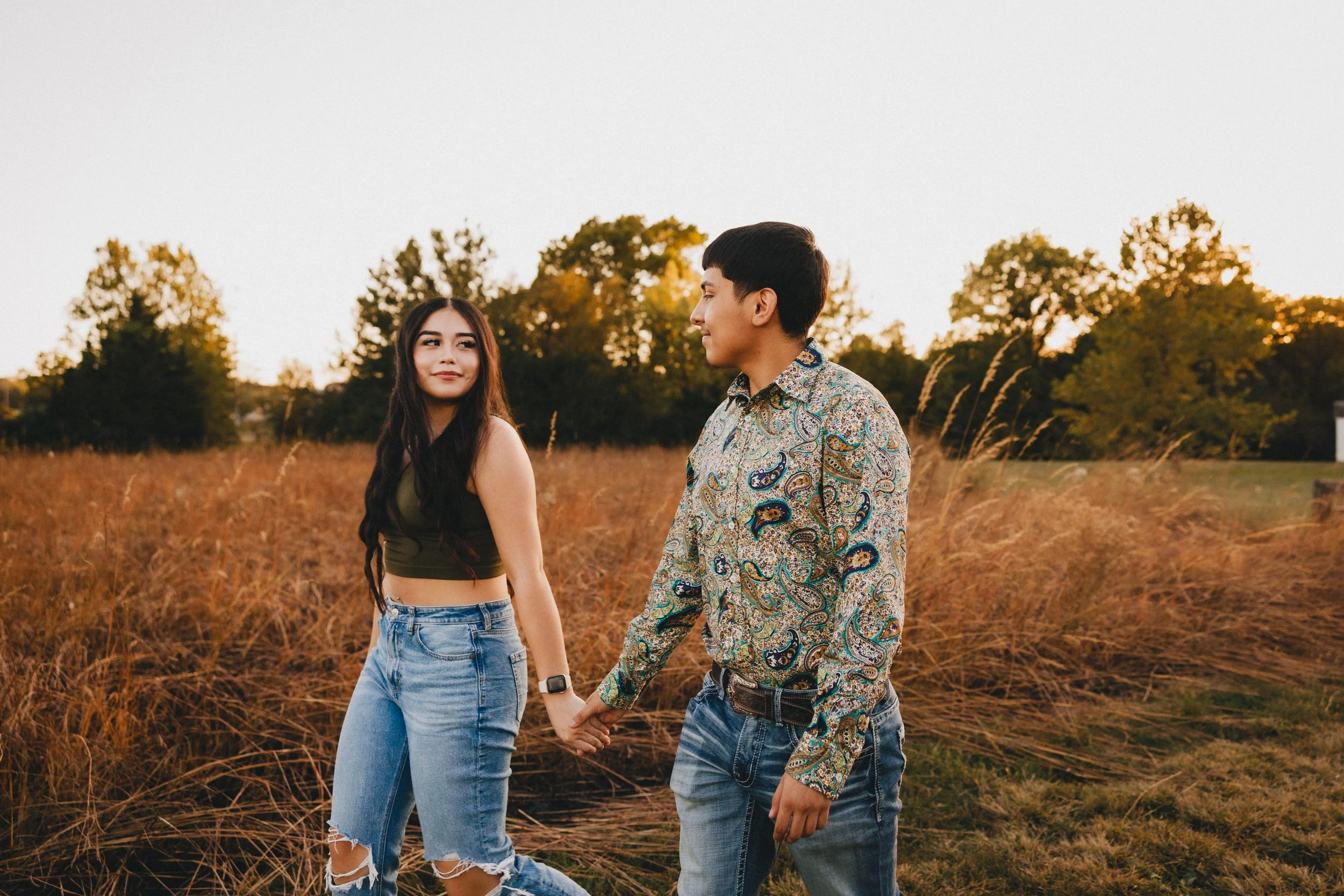 A young woman and man walking hand-in-hand through a field at sunset, with trees in the background. Shot in Topeka, Kansas. 
