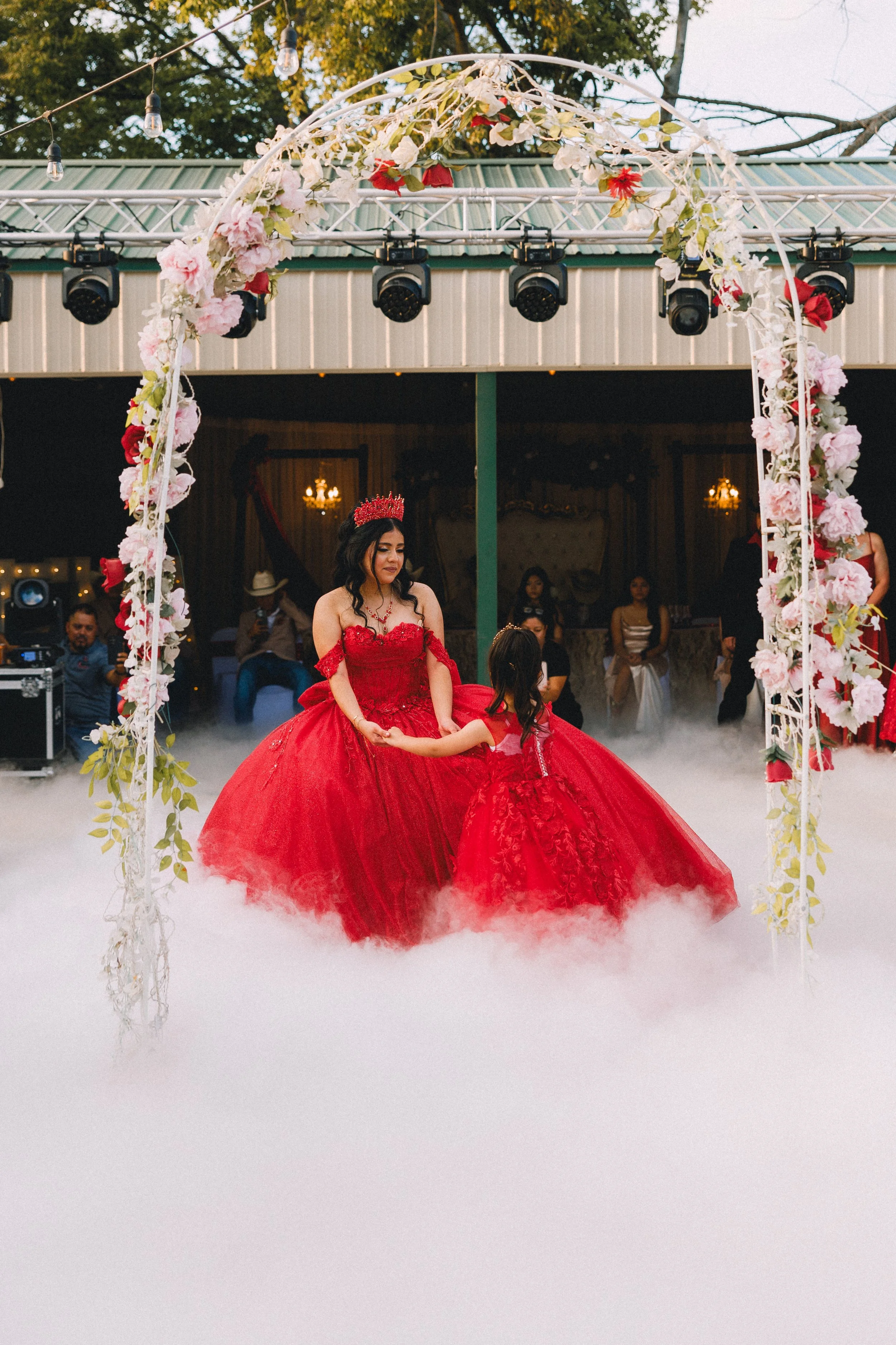 A woman and a young girl are dressed in matching red ball gowns, dancing under a floral arch with fog on the ground, at an outdoor event in the evening.
