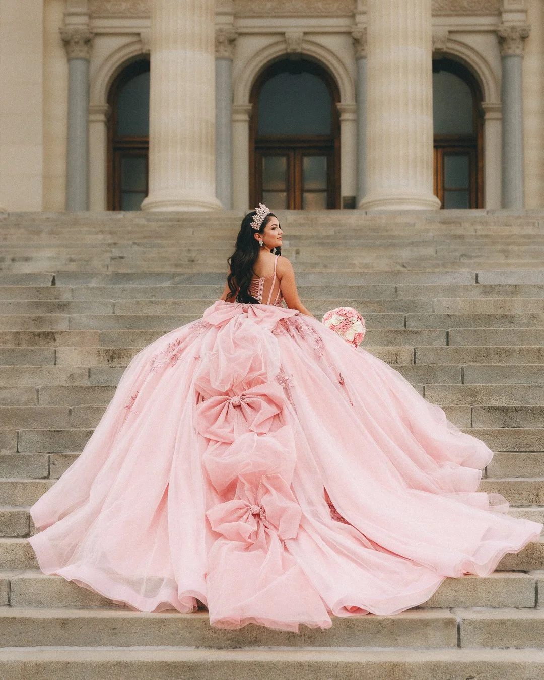 A woman in a pink ball gown with large bows sits on the steps of a neoclassical building with tall columns.