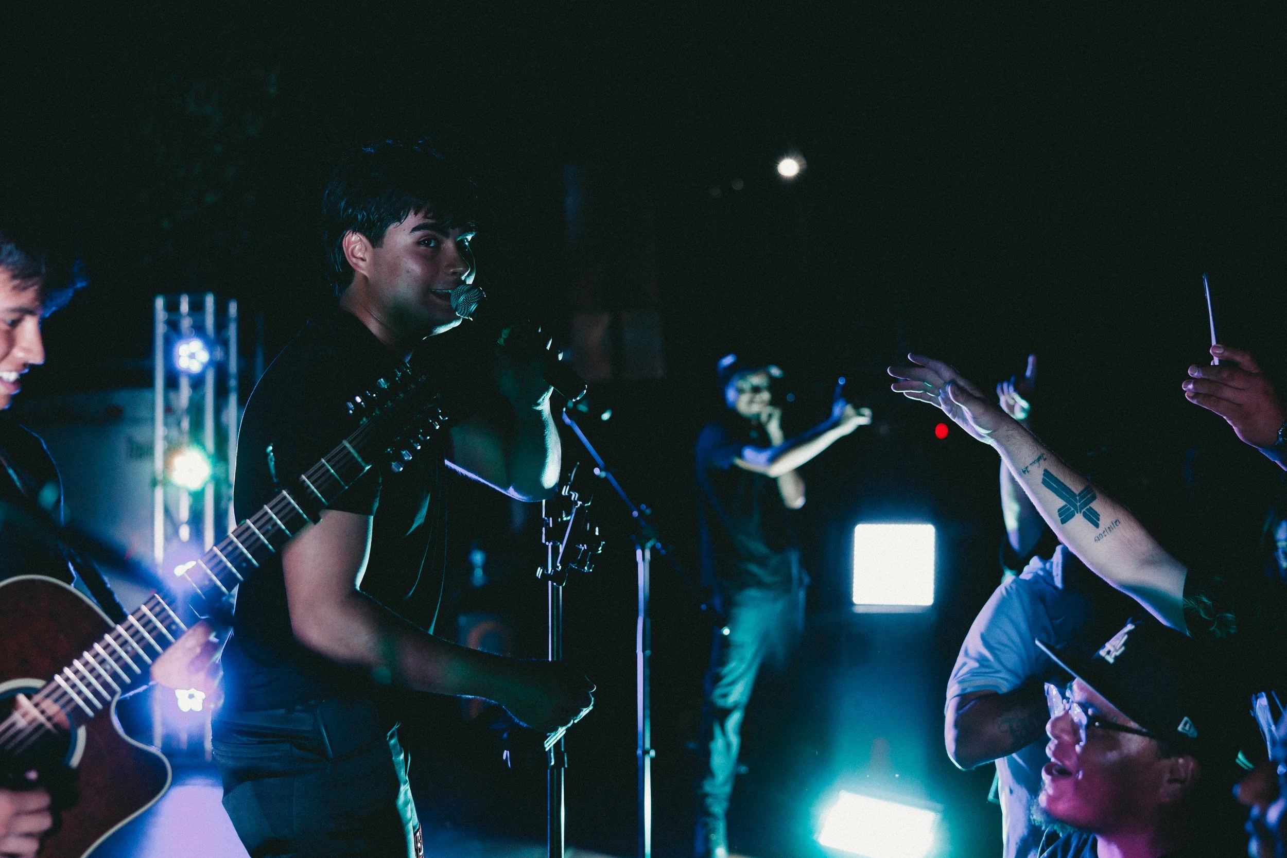 A live musical performance with a singer at the microphone, a guitarist, and a person in the background with arms raised, in a dark setting with bright stage lights and audience reaching out. Shot at Fiesta Topeka.