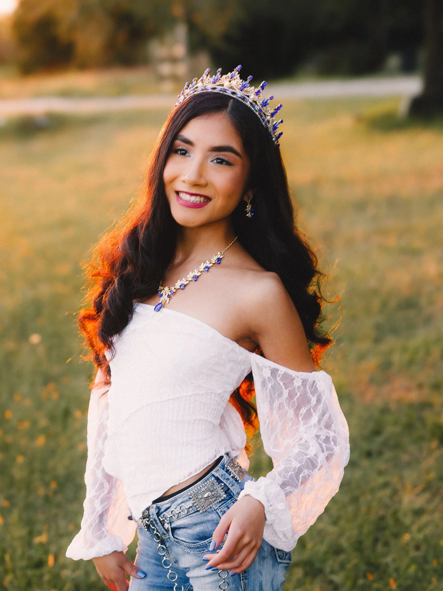 A young woman with long dark hair, wearing a tiara, a white off-shoulder lace top, and blue jeans, stands in a park during sunset, smiling.