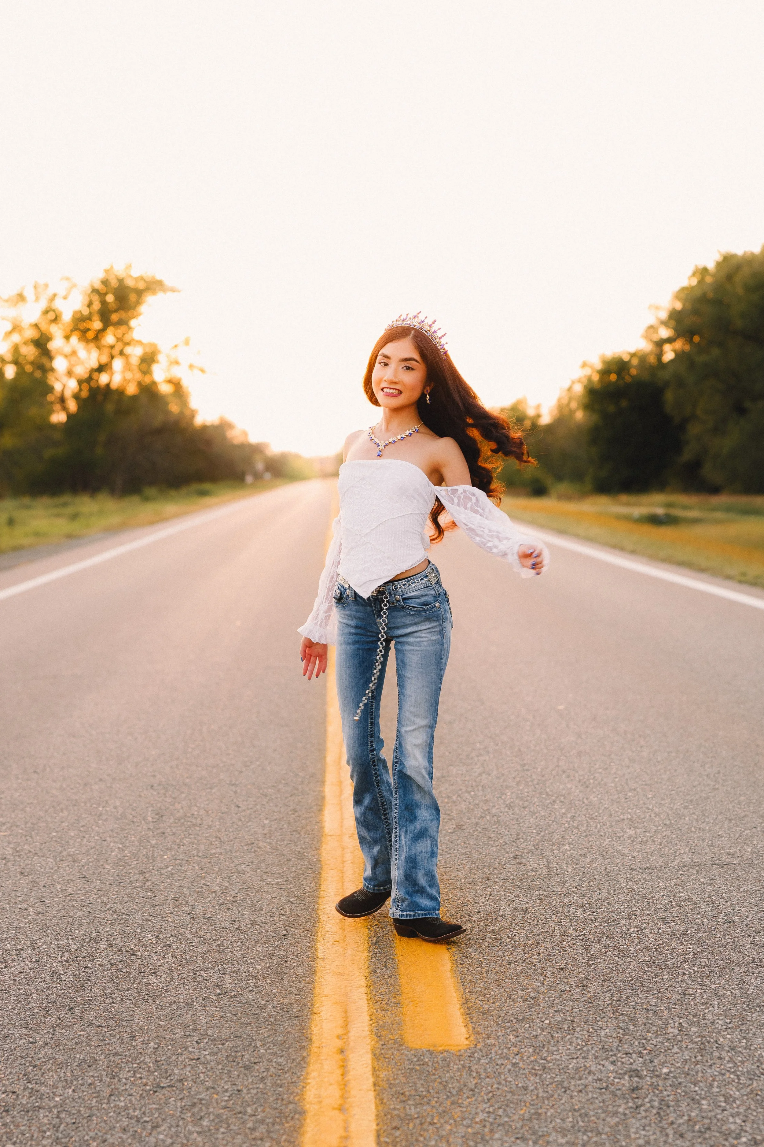 A young woman wearing a crown, necklace, and earrings, standing in the middle of a road at sunset, smiling and posing with one arm extended.