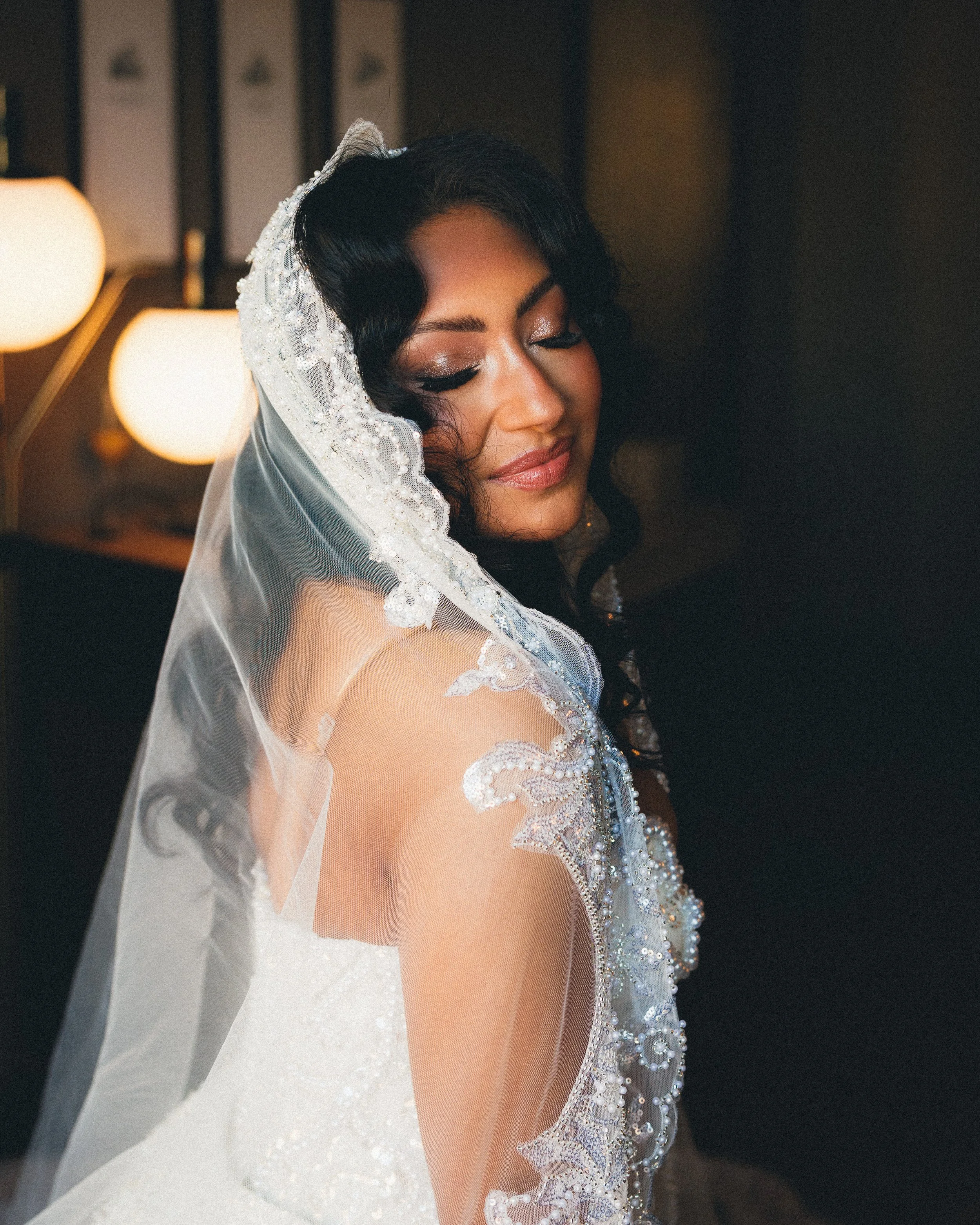 Portrait of a bride with dark curly hair and a lace veil, smiling softly with closed eyes, indoors with warm lighting. Shot at Cyrus Hotel in Topeka, Kansas.