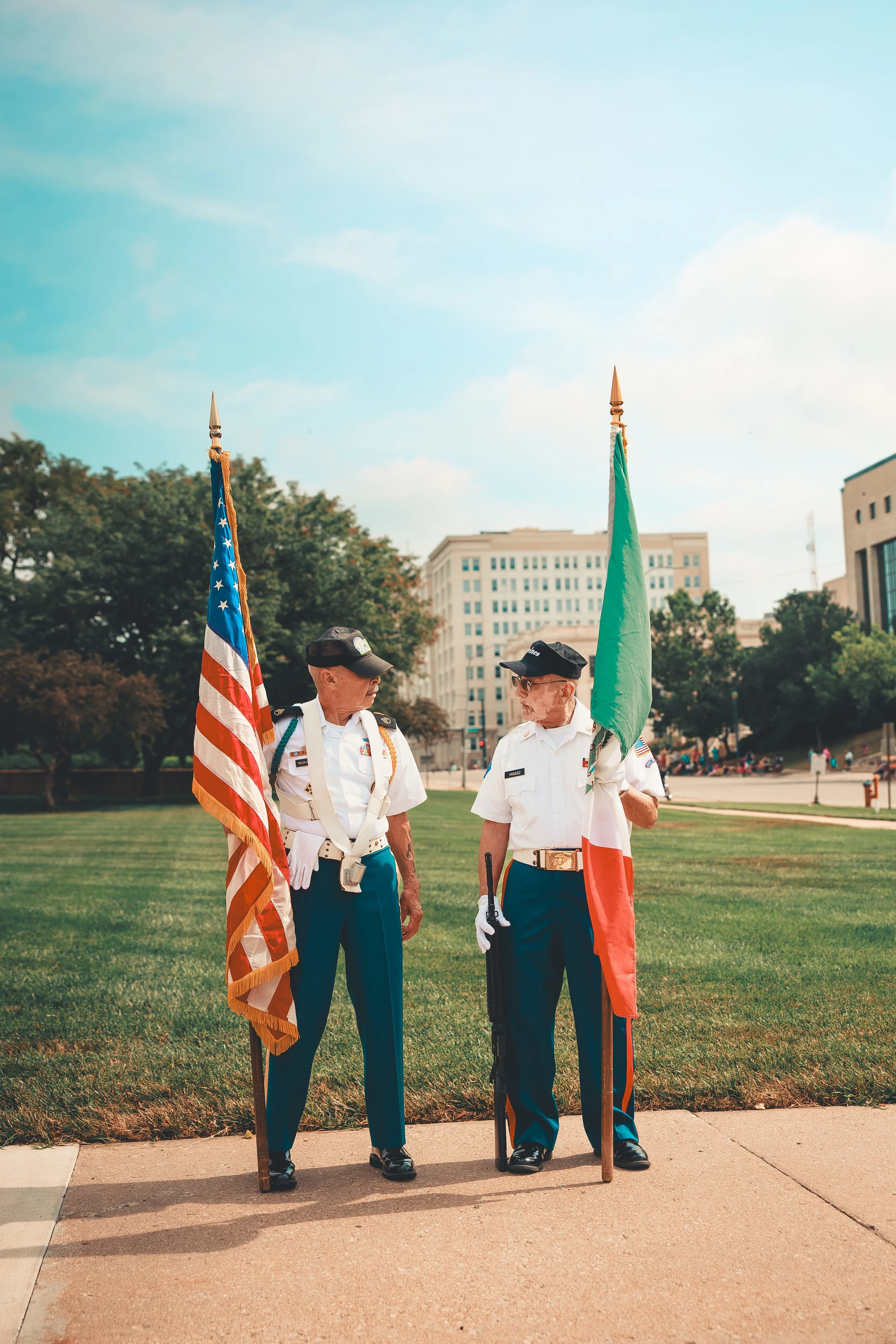 Two uniformed veterans standing outdoors, holding the United States and Italian flags, in a park with buildings and trees in the background. Shot at Fiesta Topeka parade.