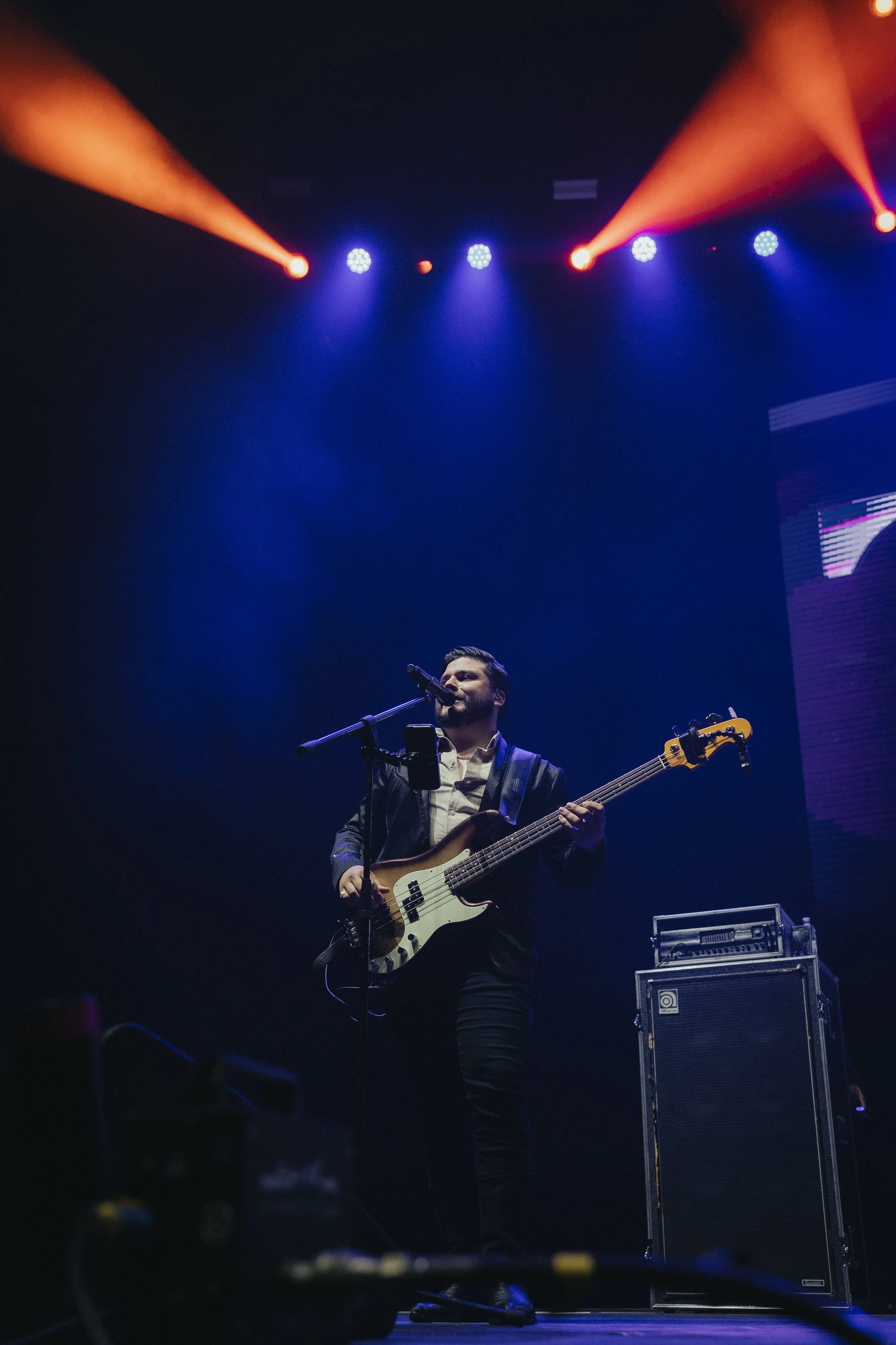 Musician on stage playing bass guitar and singing into a microphone with colorful stage lights overhead. Stormont Vail Event Center Topeka, Kansas. 