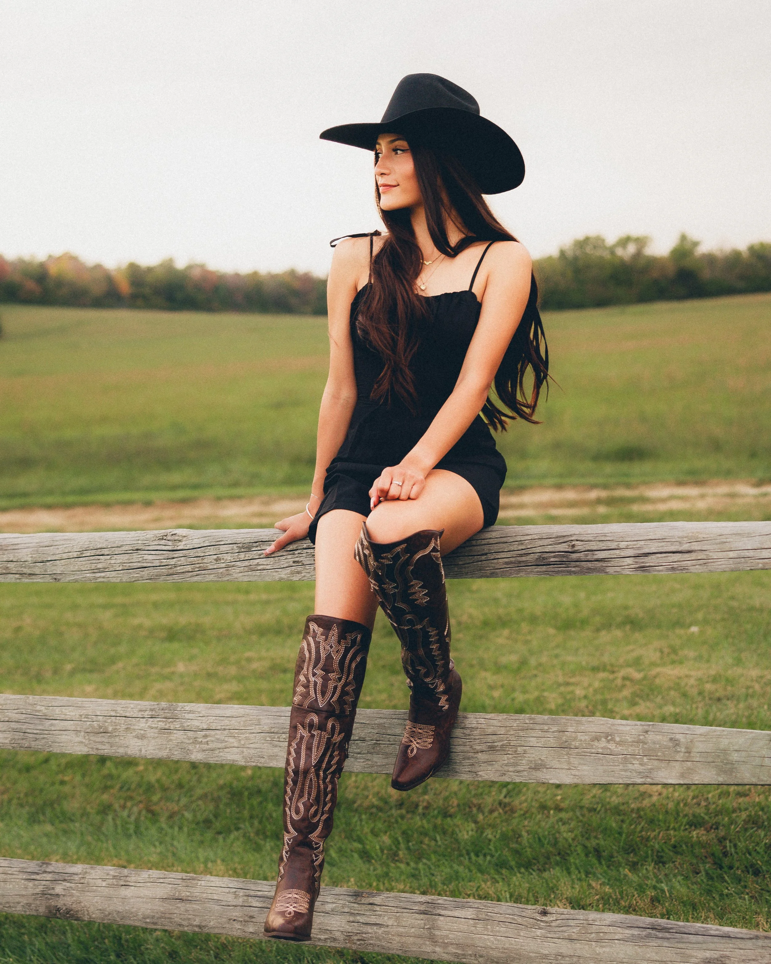 A woman wearing a large black hat, black dress, and cowboy boots sitting on a wooden fence in a grassy field.