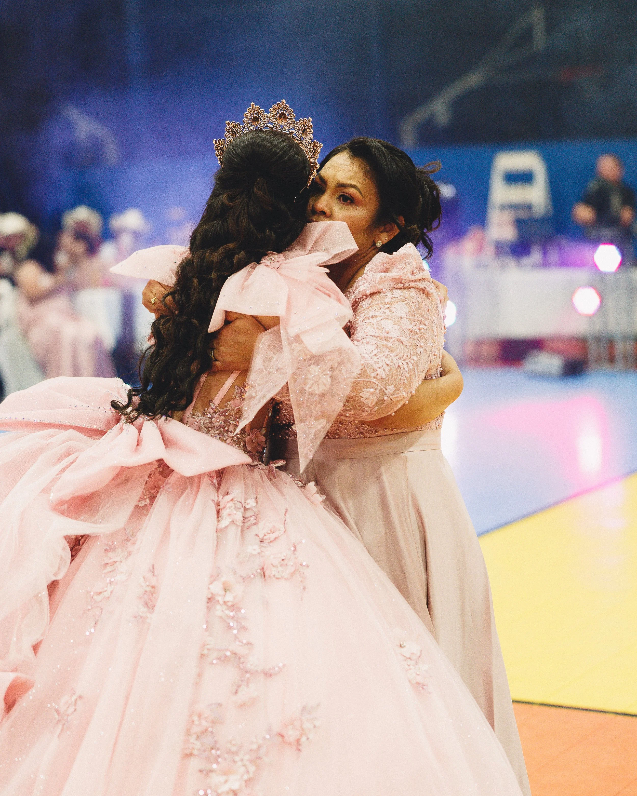 Two women embrace, one wearing a pink gown with intricate details and a tiara, and the other in a light-colored dress, at a celebration or event.