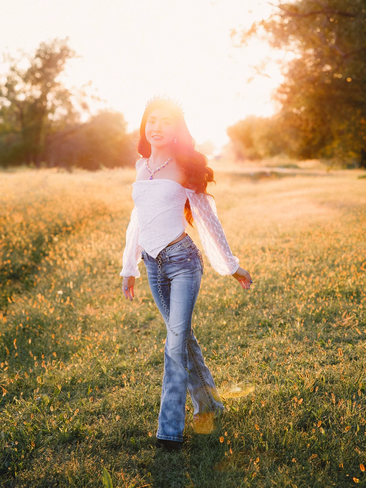 A woman with long, wavy hair standing in a field during sunset, dressed in a white off-shoulder top and jeans, smiling at the camera.
